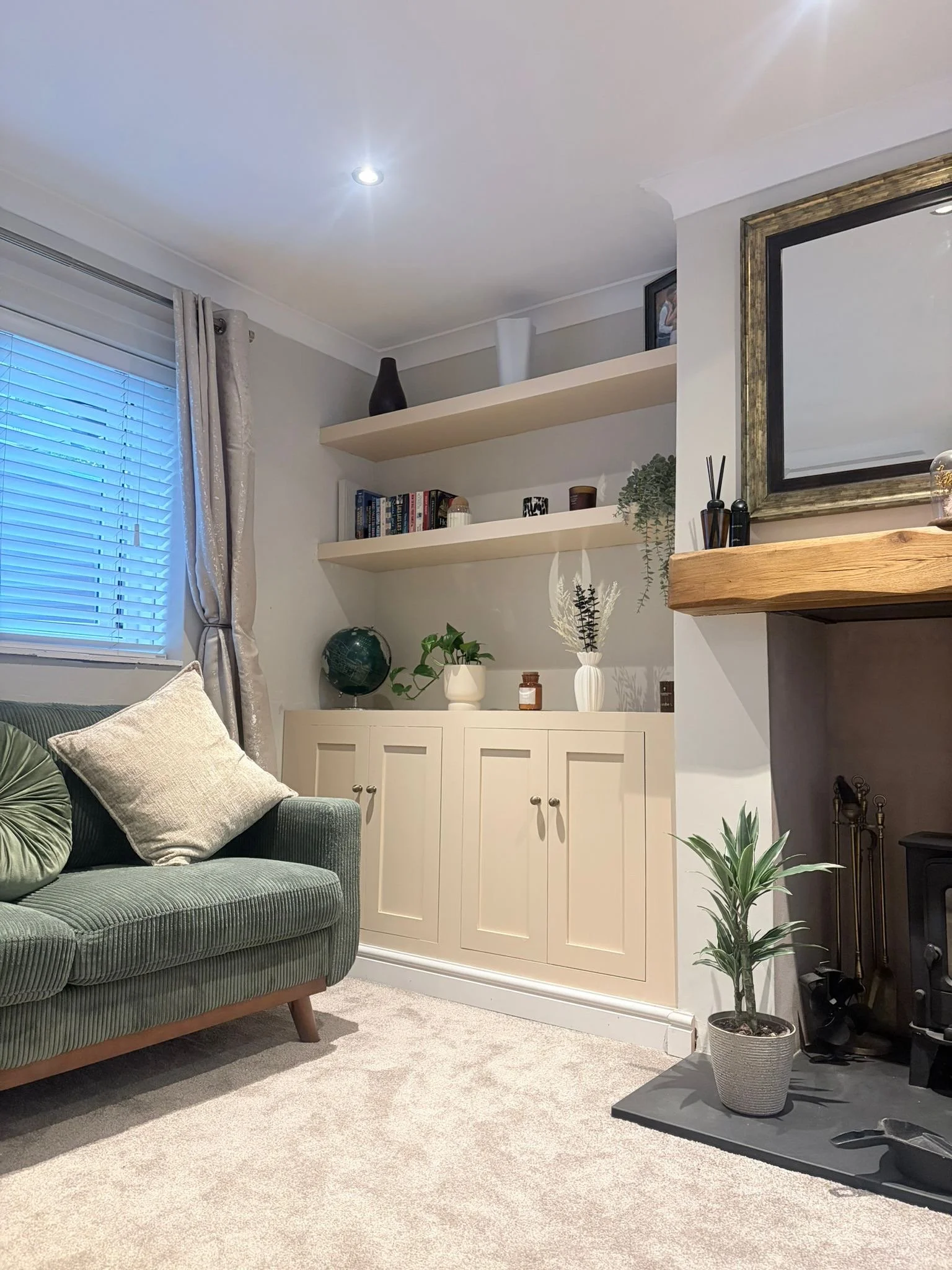 Living room with a green sofa, beige carpet, white cabinet with plants and books, a fireplace with a large mirror above, and decorative items on shelves.
