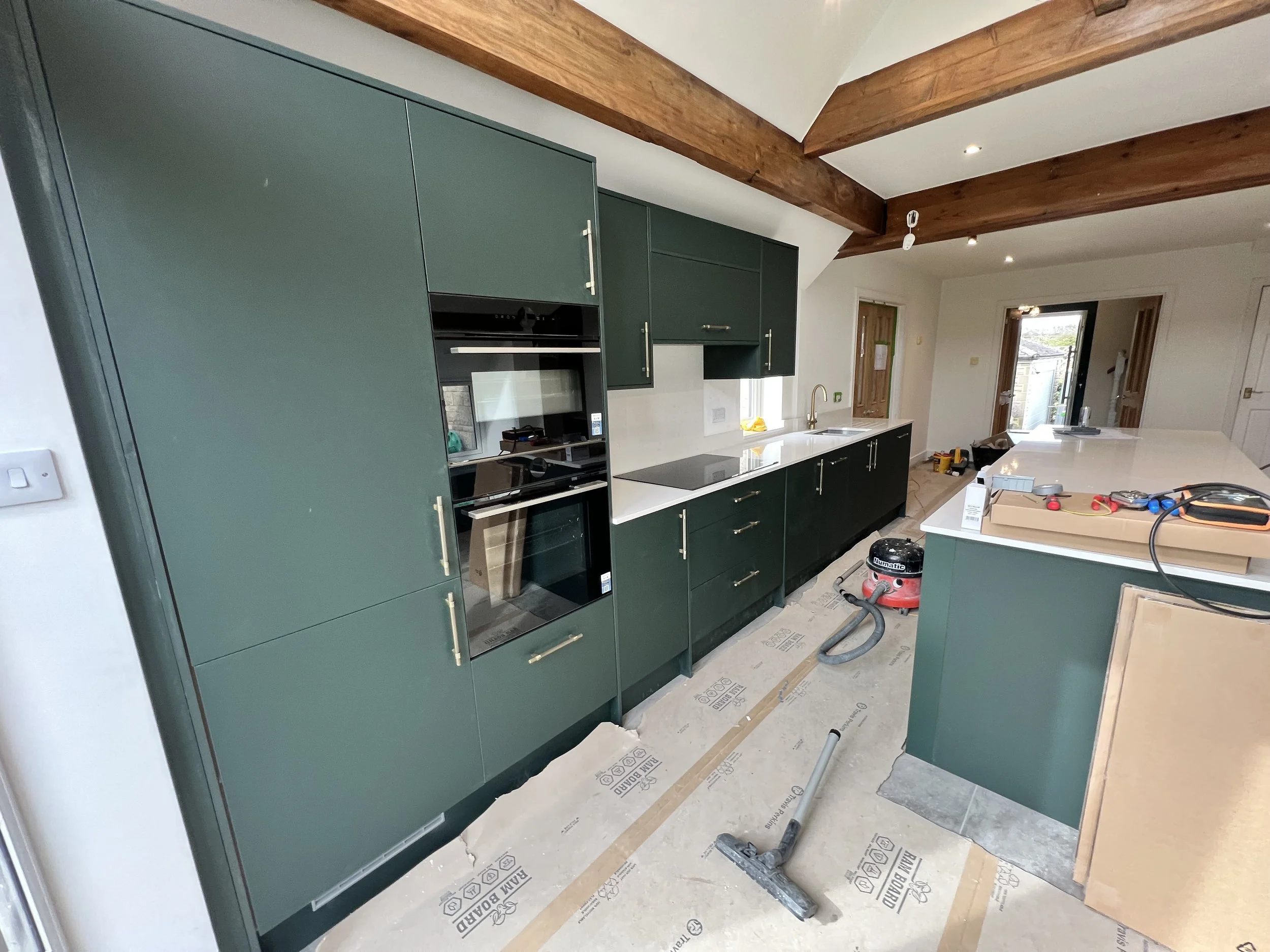 A kitchen under construction with green cabinets, a white countertop, a built-in oven, and exposed wooden ceiling beams. Construction tools and materials are scattered on the floor.