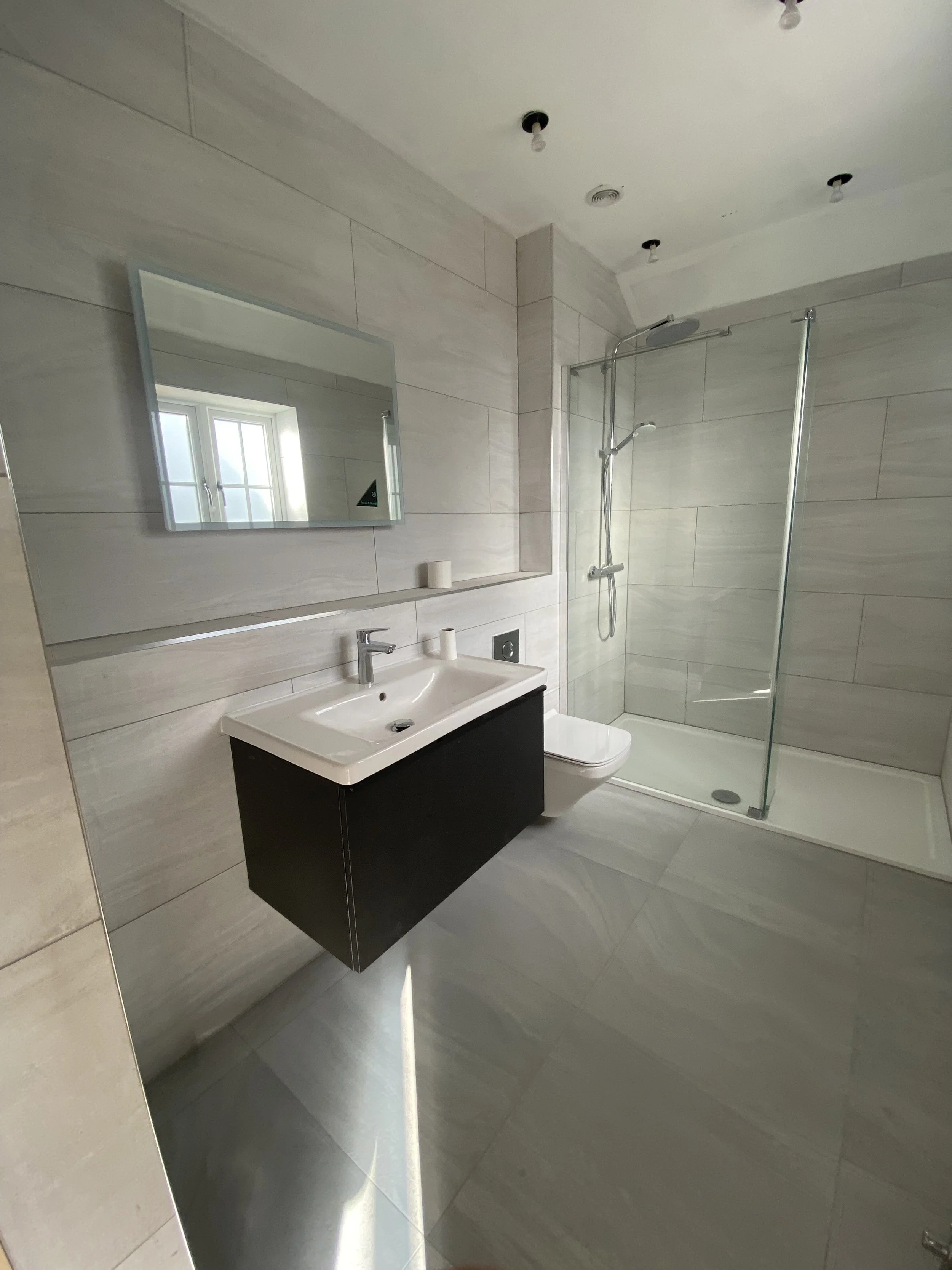 Modern bathroom with large mirror, white sink with black vanity, wall-mounted toilet, walk-in shower with glass enclosure, and beige tiled walls and floor.