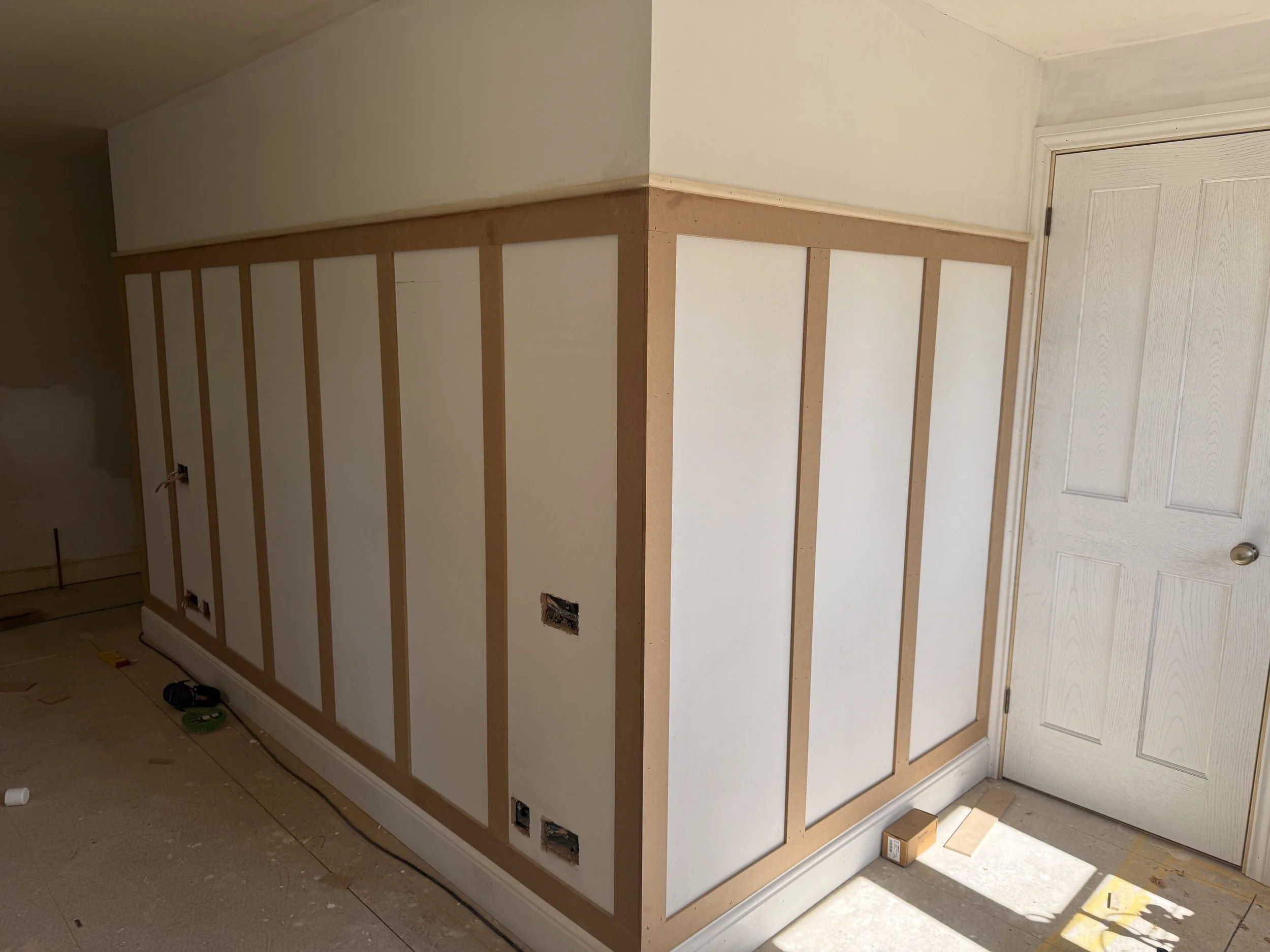 Partially constructed wall with vertical wooden studs and white drywall, next to a white door with an unfinished floor.