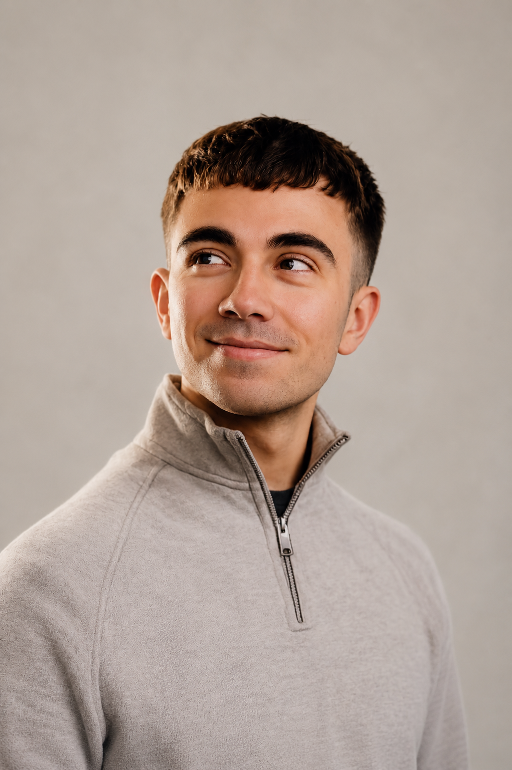 A young man with dark hair and light skin, smiling slightly, wearing a beige quarter-zip sweater, looking to his left, standing against a gray background.