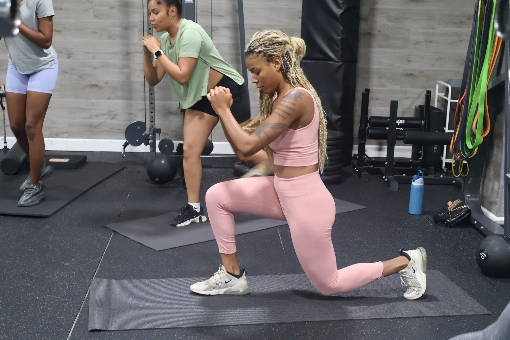A woman with long dreadlocks in pink workout clothes including a sports bra and leggings is performing a kneeling lunge exercise on a gym mat, with her hands clasped together in front of her chest. Behind her, two women are standing: one in gray shor