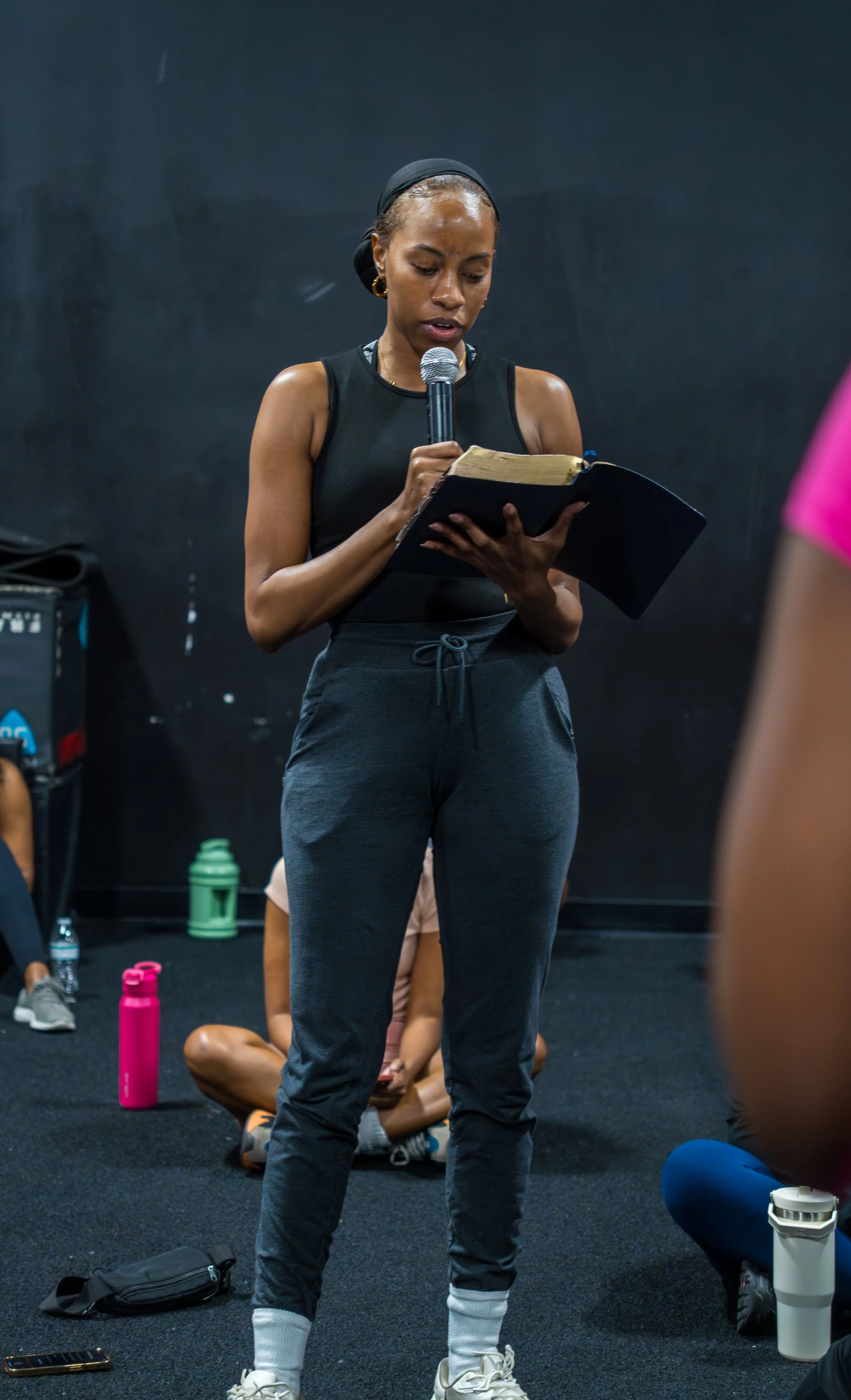 A woman in athletic clothing reading from a book while holding a microphone in a fitness studio.