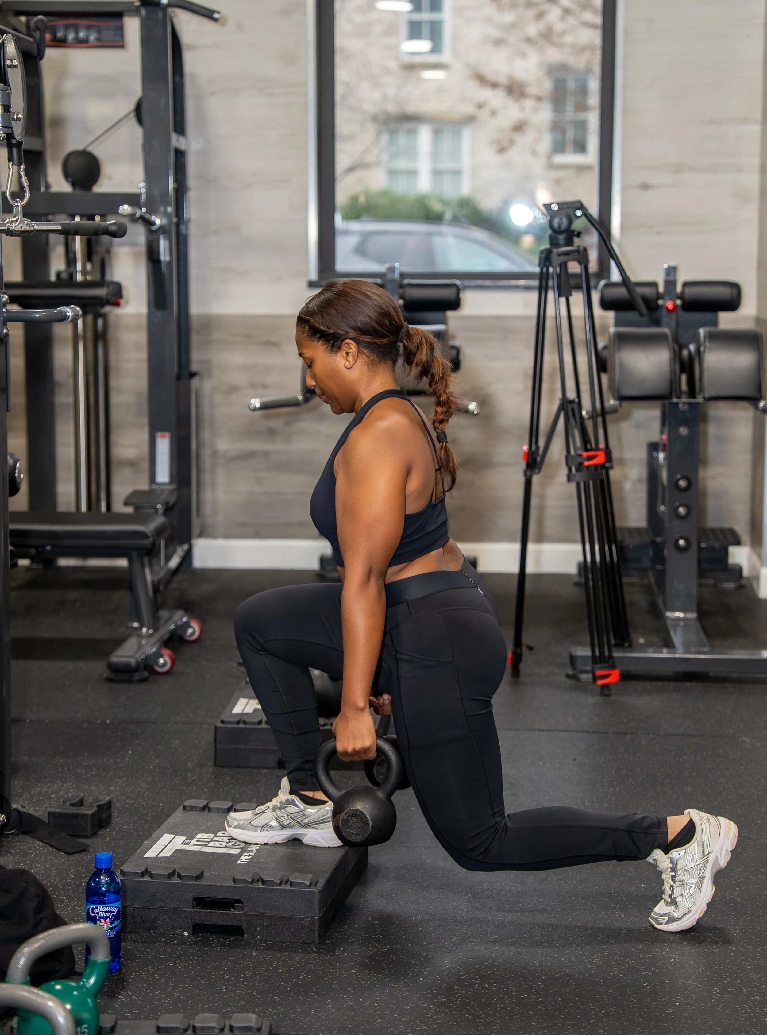 A woman in workout clothes performing a kneeling exercise with a kettlebell in a gym with workout equipment and a tripod with camera nearby.
