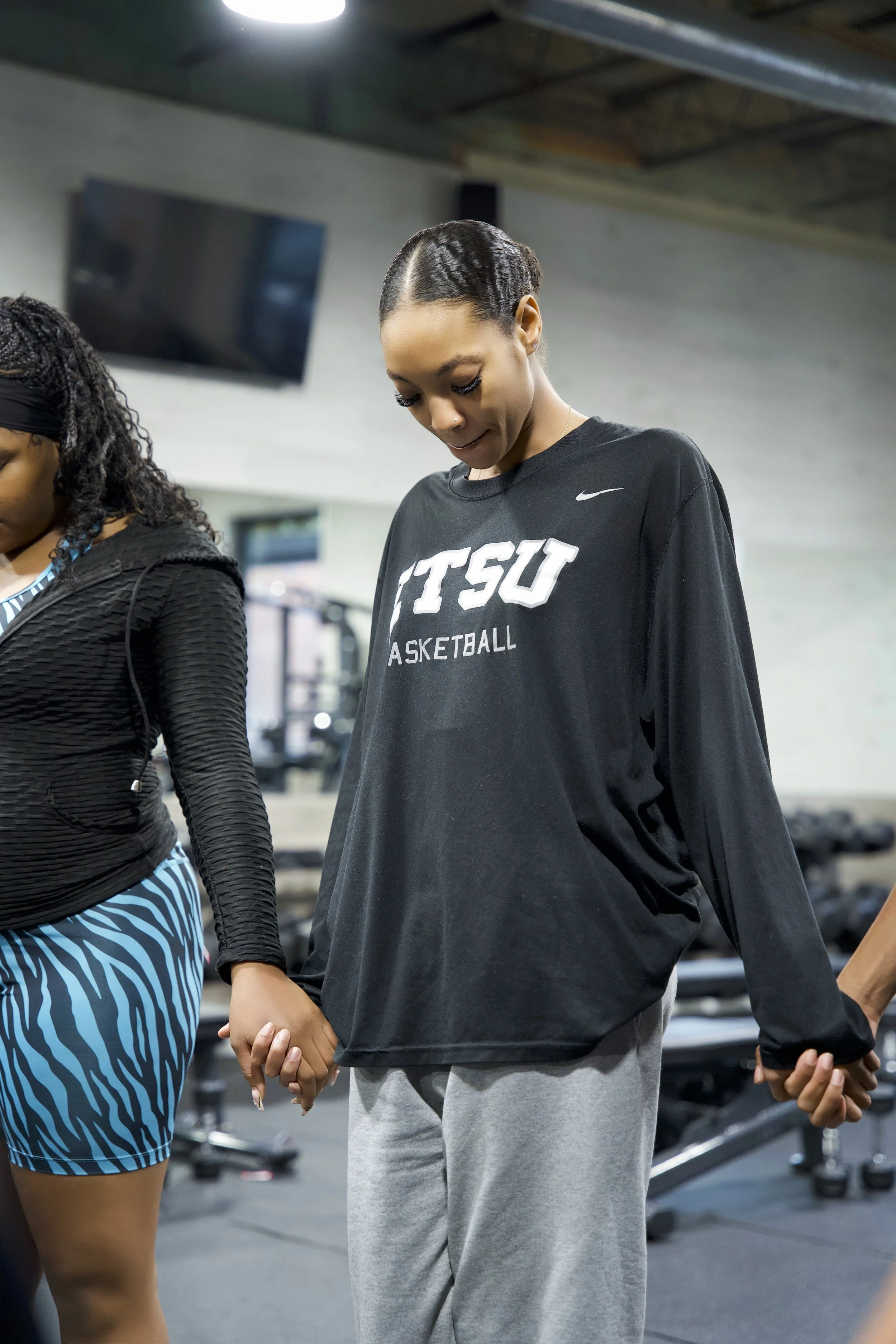 Two women holding hands in a gym. One woman is wearing a black TTSU basketball shirt and gray sweatpants. The other woman, partially visible, is wearing a black jacket and blue striped shorts.