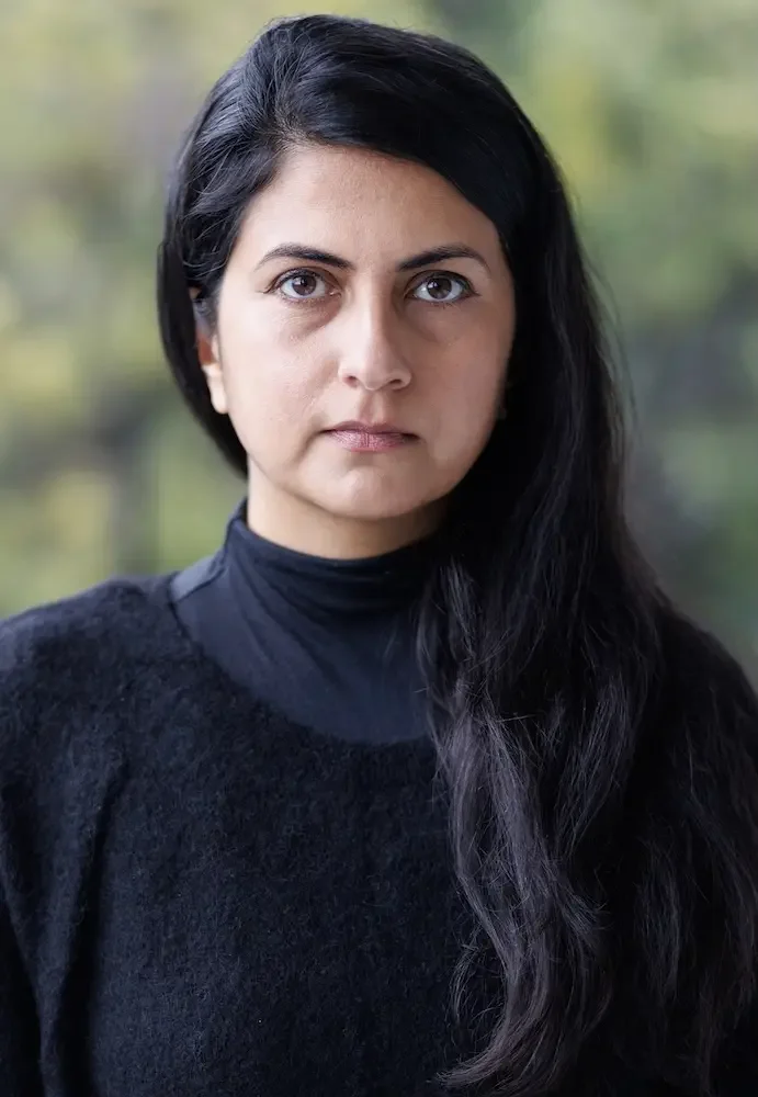 A woman with long dark hair and fair skin, wearing a black sweater and standing outdoors with a blurred background of trees.
