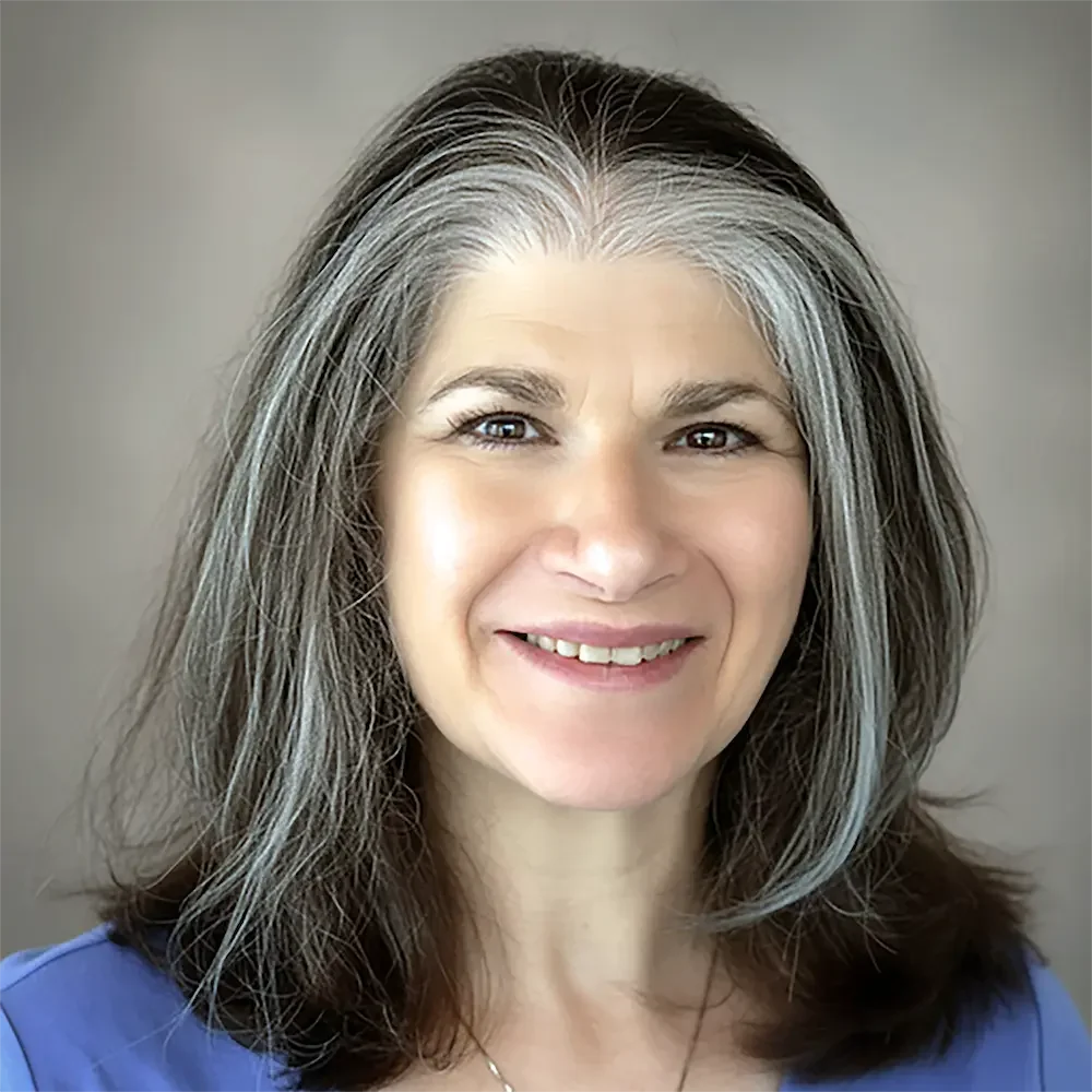 A woman with shoulder-length brown and gray hair, smiling, wearing a blue top, against a neutral background.