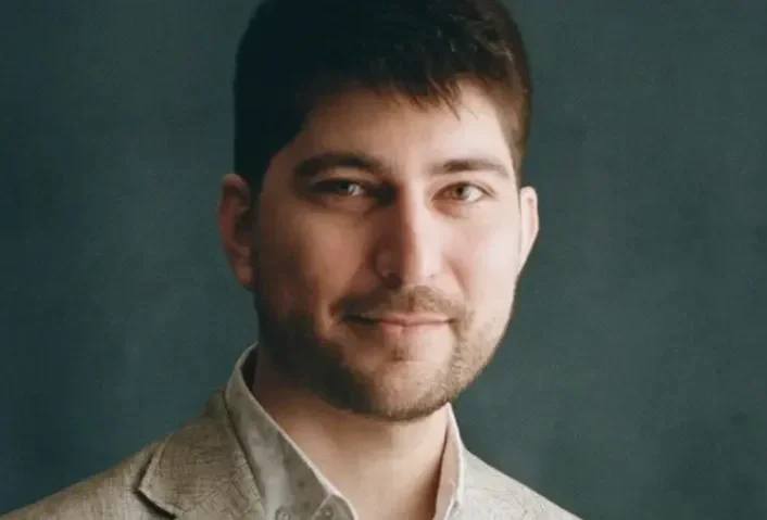 A portrait of a young man with dark hair, wearing a beige blazer over a light-colored shirt, smiling softly against a dark backdrop.
