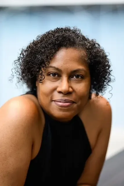 A woman with curly hair looking at the camera with a slight smile, wearing a black sleeveless top.