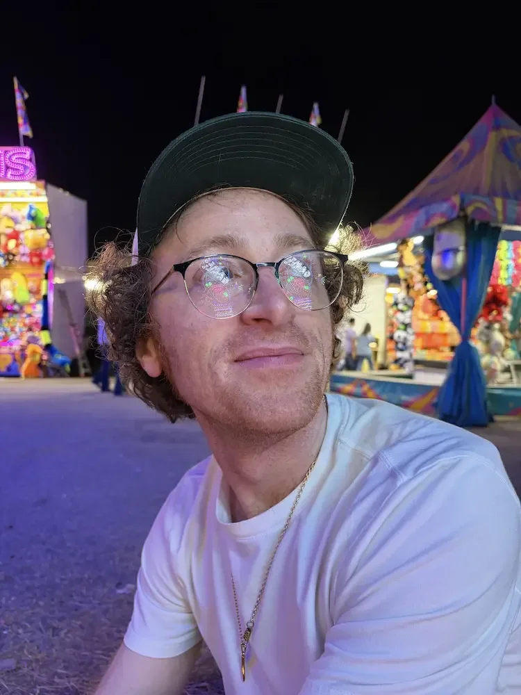 A young man with curly hair, glasses, and a black baseball cap, sitting at a carnival at night. Bright, colorful games and tents are visible in the background.