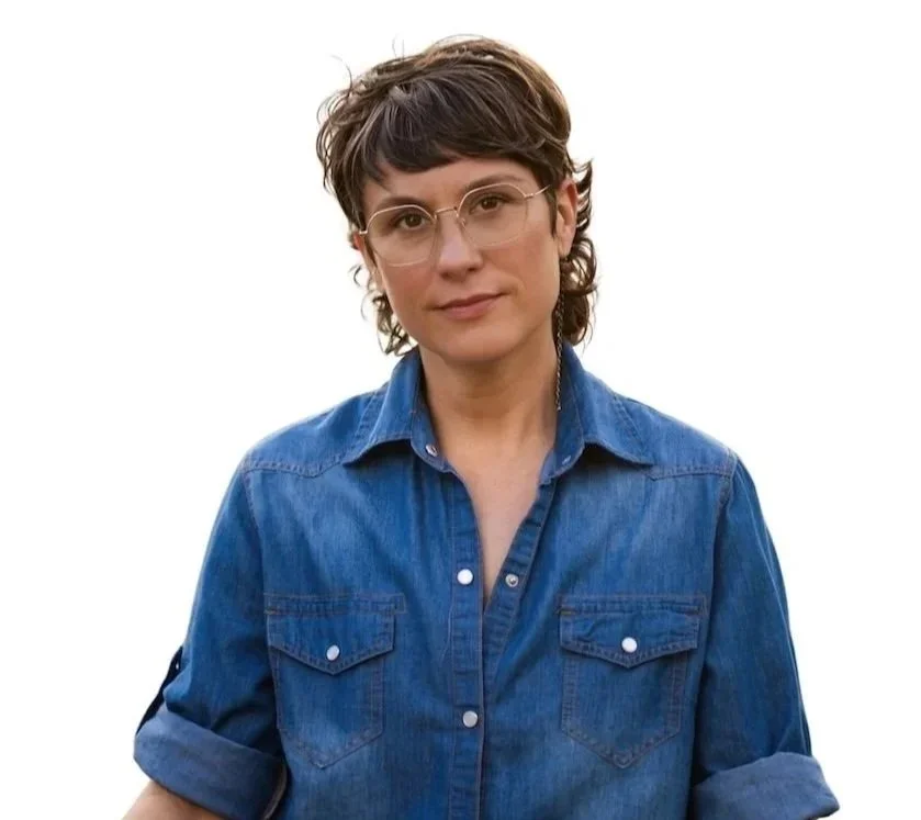 A woman with short brown hair wearing glasses and a blue denim shirt thoughtfully looking at the camera.
