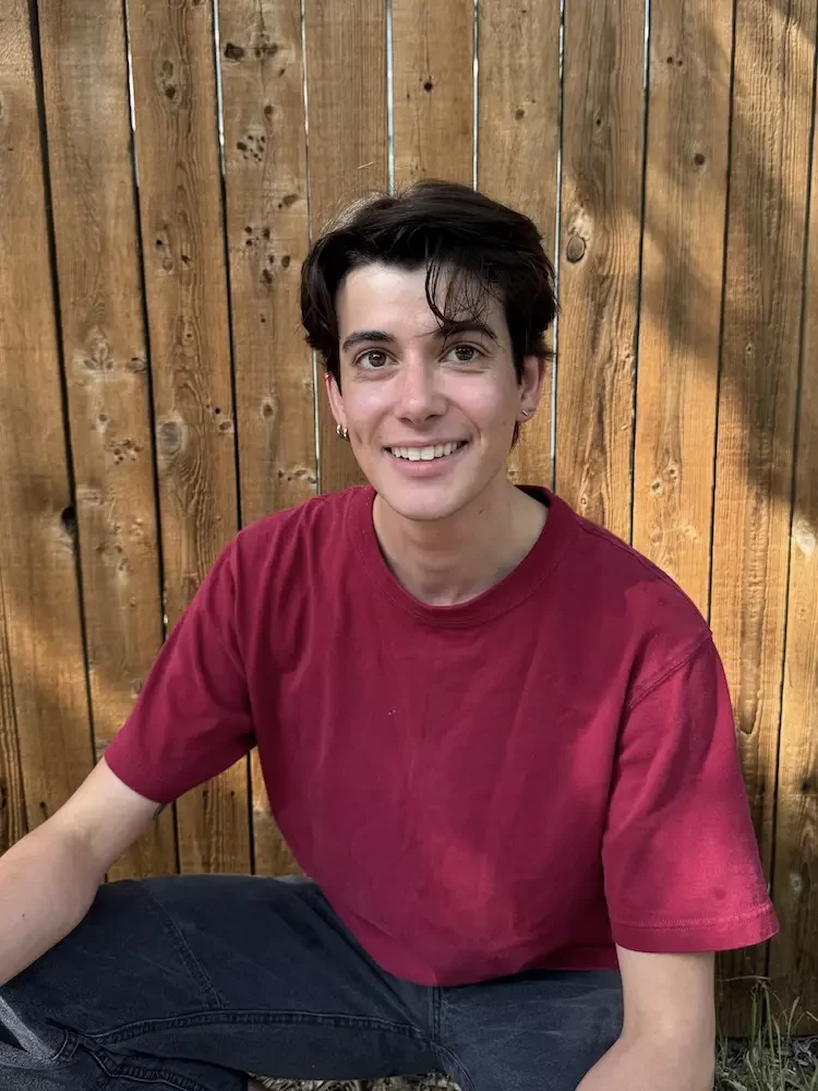 A young man with dark hair, wearing a red t-shirt, sitting outside against a wooden fence and smiling at the camera.