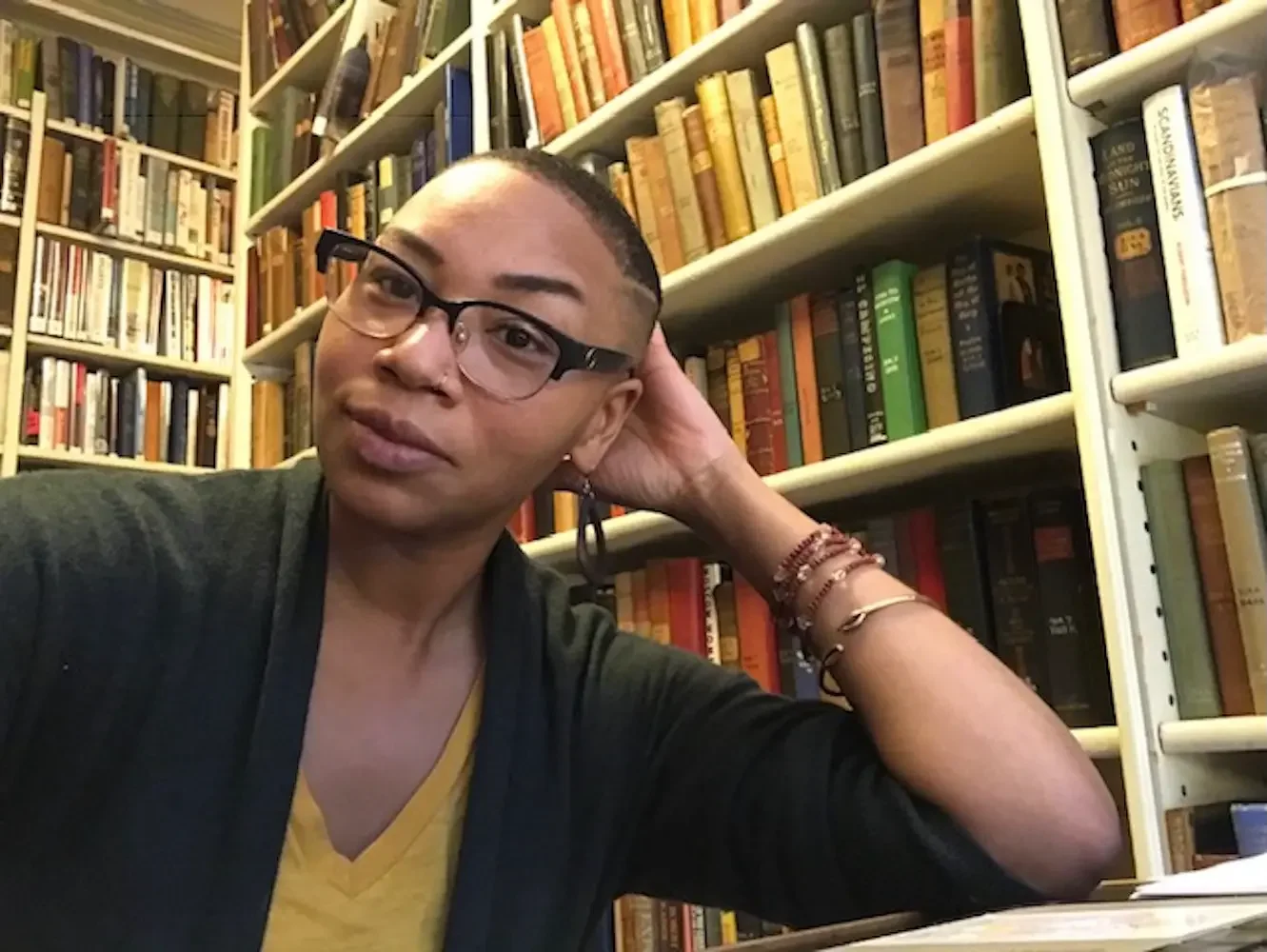 A person with short hair and glasses sitting in front of a bookshelf filled with colorful books, resting their head on their hand, wearing a yellow shirt and bracelets.