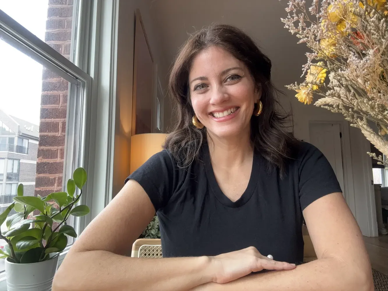 A woman with dark brown hair, wearing a black t-shirt and gold hoop earrings, smiling while sitting at a table by a window with a potted plant and dried flowers in the background.