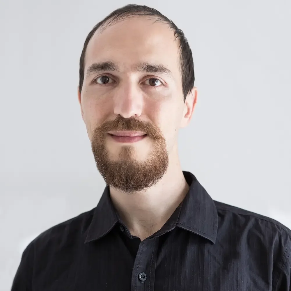 Portrait of a man with a beard and mustache wearing a black collared shirt against a light gray background.