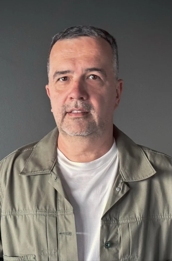 A middle-aged man with short dark hair, some gray, and a light beard, wearing a beige jacket over a white T-shirt, standing against a plain gray background.