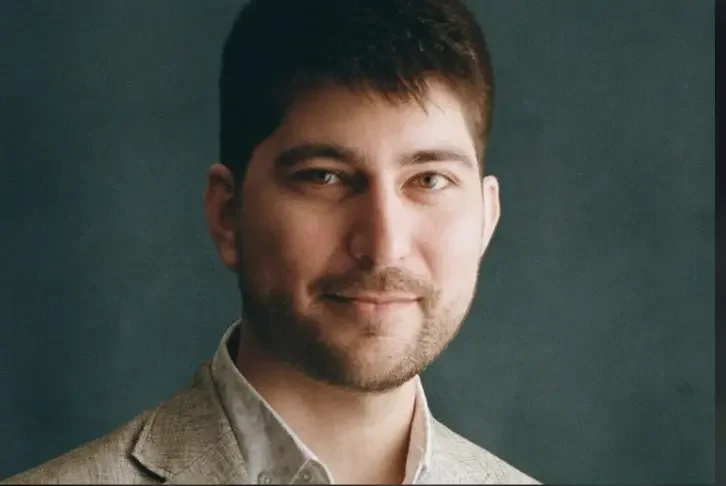 Portrait of a young man with short dark hair, wearing a light-colored blazer and a white shirt, against a dark background.