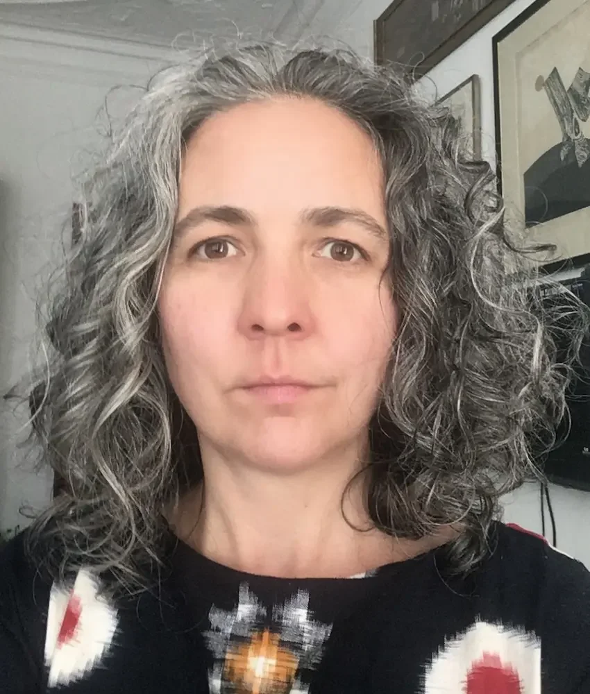 A woman with shoulder-length curly gray hair looking directly at the camera, wearing a black top with a colorful pattern, in a room with framed pictures on the wall.