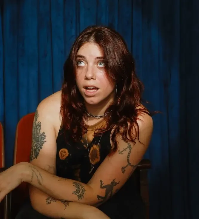 Young woman with curly red hair and tattoos on her arms, sitting against a blue curtain background, looking upward with an expressive face.