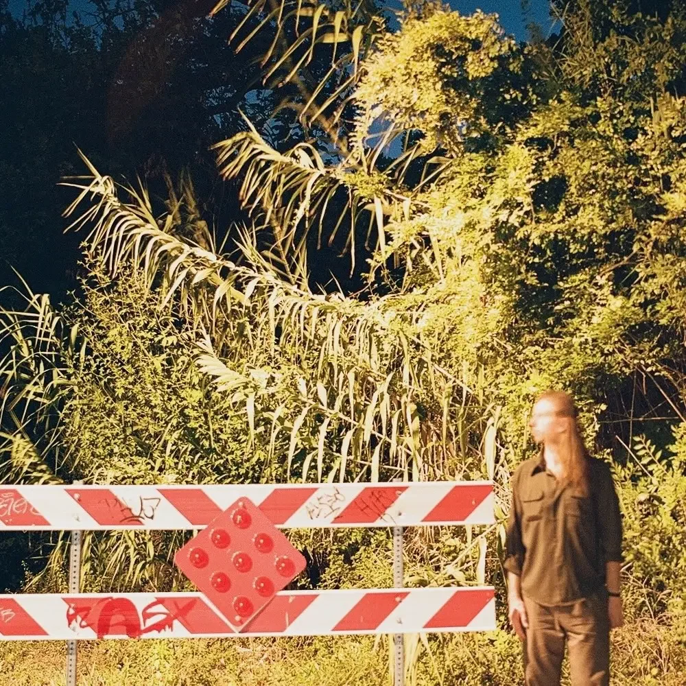 Nighttime scene of a woman in a brown jacket standing next to a red and white barricade with an illuminated lush green foliage background.