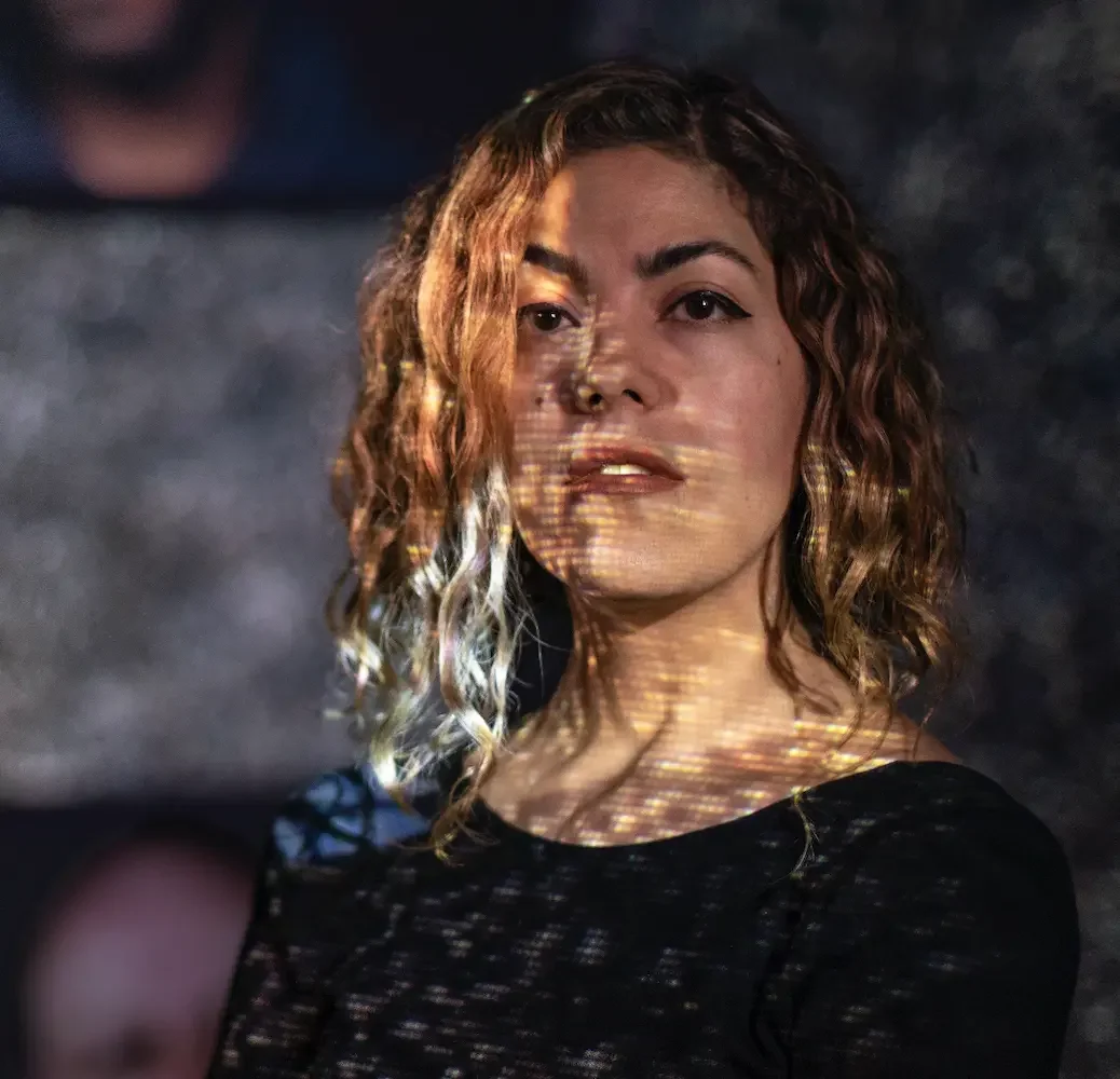 A woman with curly blonde hair looking at the camera with projected light patterns on her face and chest, wearing a black top.