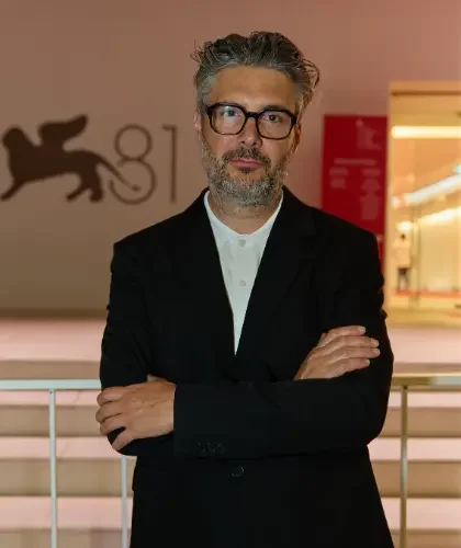 Man with gray hair, beard, and glasses standing with arms crossed in front of a red background and a lion logo.