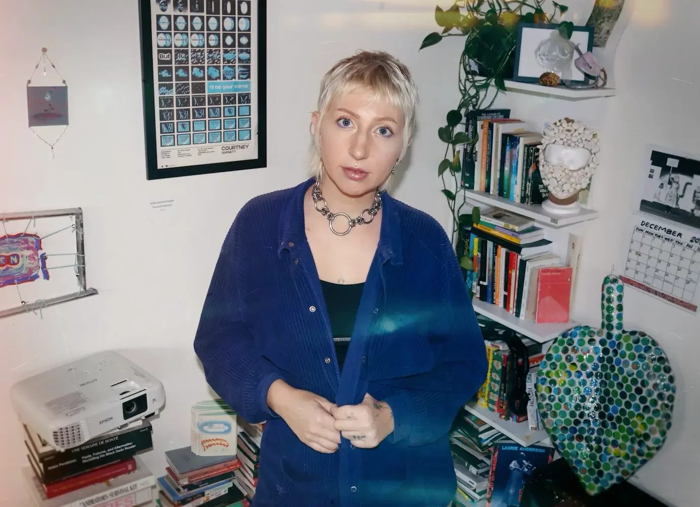 A woman with short blonde hair and blue eyes standing in a cluttered room, wearing a blue jacket and a silver chain necklace. The room has bookshelves, a heart-shaped mosaic art piece, and various decorative items.