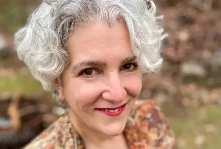 A woman with short, curly gray hair smiling outdoors, with a blurred background of trees and natural surroundings.