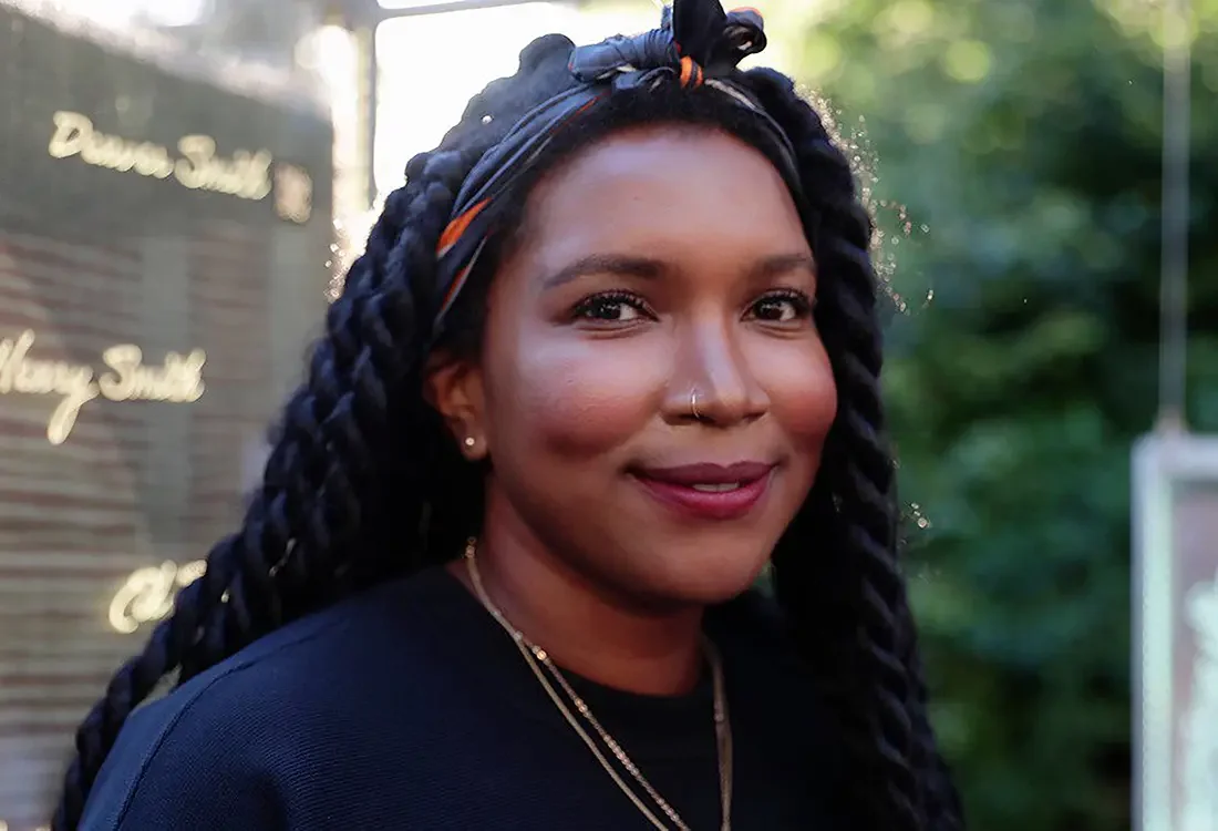 A woman with braided hair, wearing a headband, earrings, a nose ring, and layered necklaces, smiling outdoors.