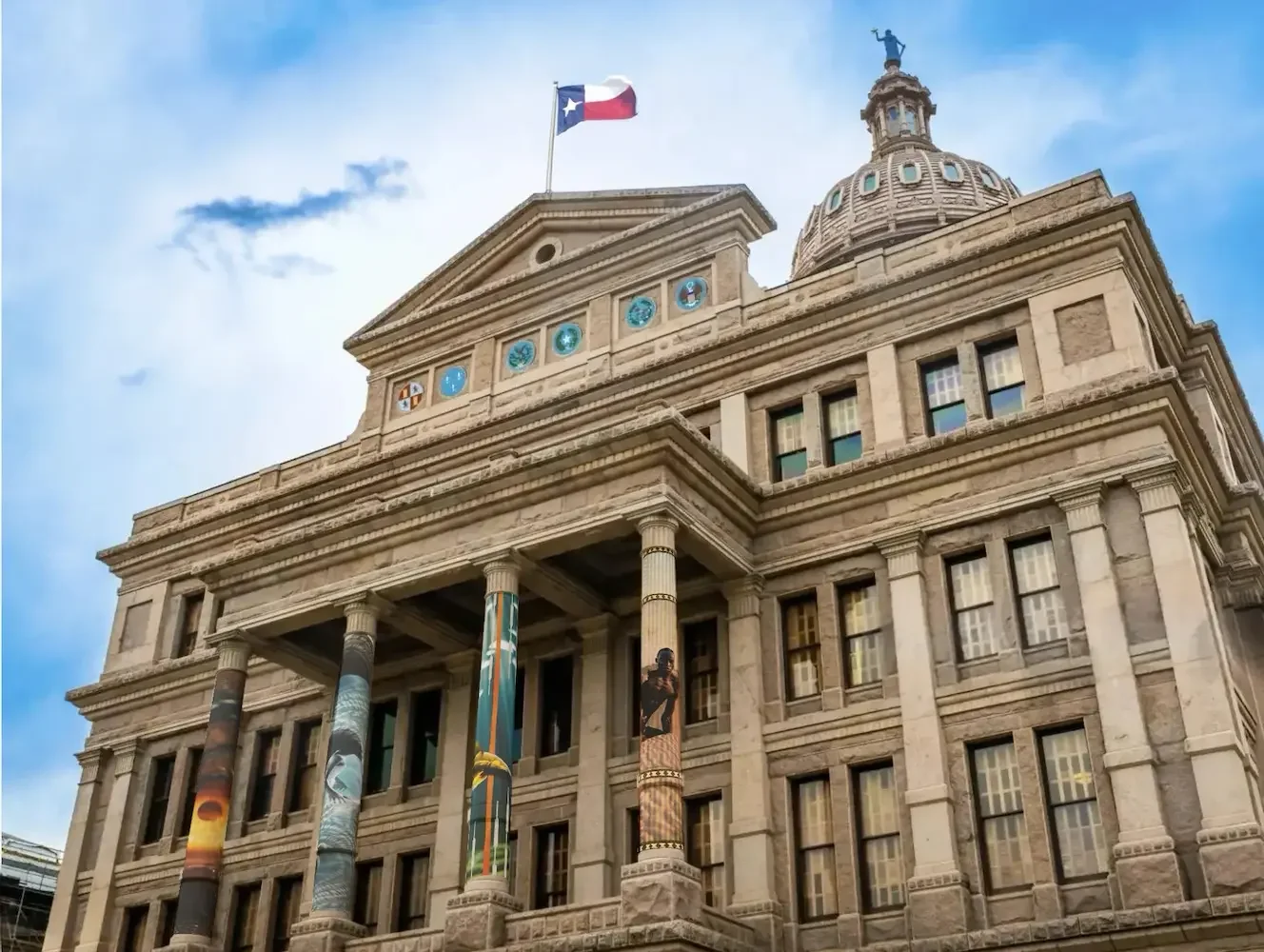 The Texas State Capitol building with a Texas flag on top, columns, and a rounded dome with a statue on top, under a blue sky with a few clouds.