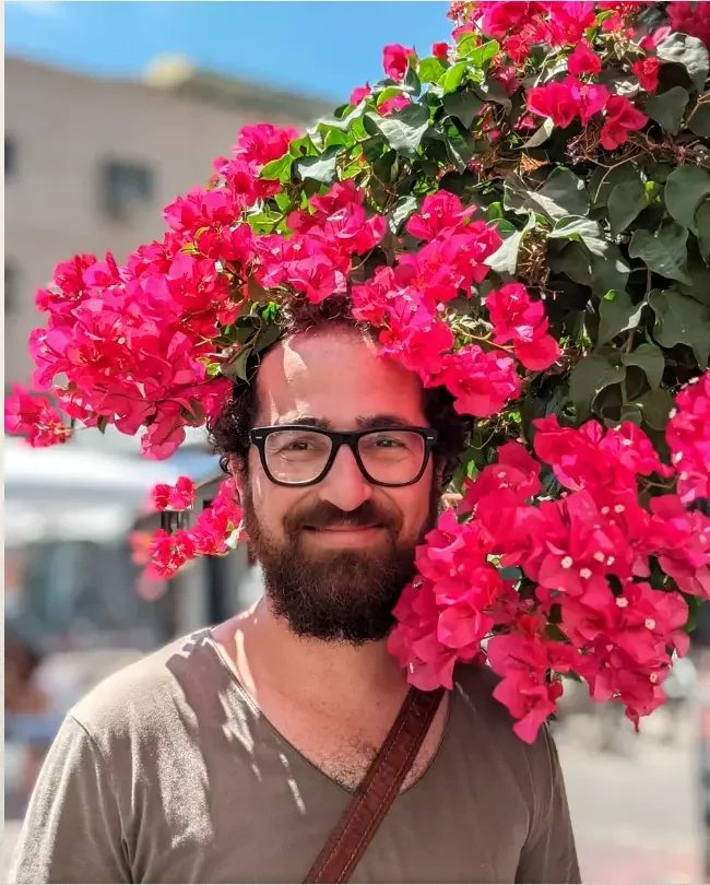 A man with glasses and a beard standing outdoors, surrounded by vibrant pink bougainvillea flowers on a sunny day.
