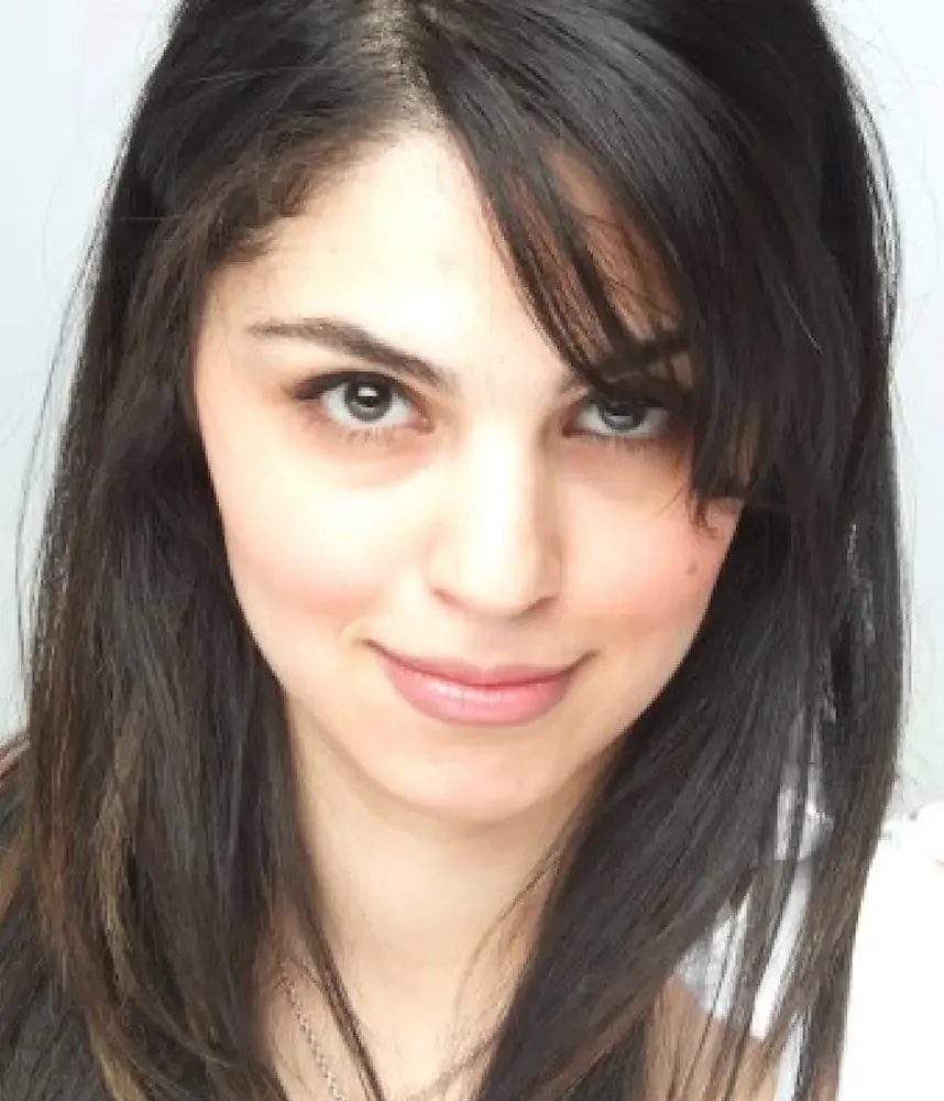 Close-up of a young woman with dark brown hair and blue eyes, smiling softly, against a plain background.