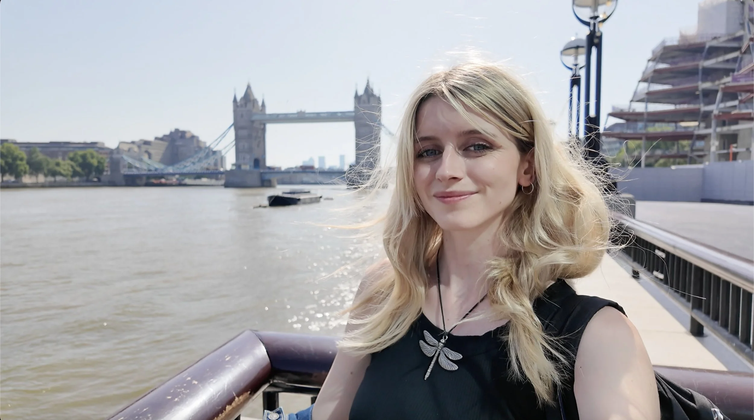 A young woman with blonde wavy hair smiling on a boat with the Tower Bridge in London visible in the background.