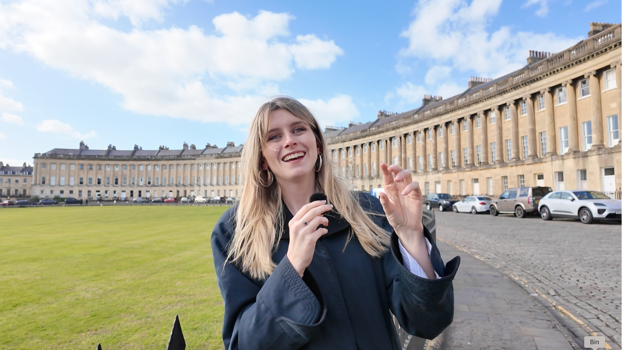 A young woman with long blonde hair, smiling and holding a microphone, standing outdoors in front of a curved row of historic stone buildings and a parking lot with several cars, on a bright day with blue sky and white clouds.