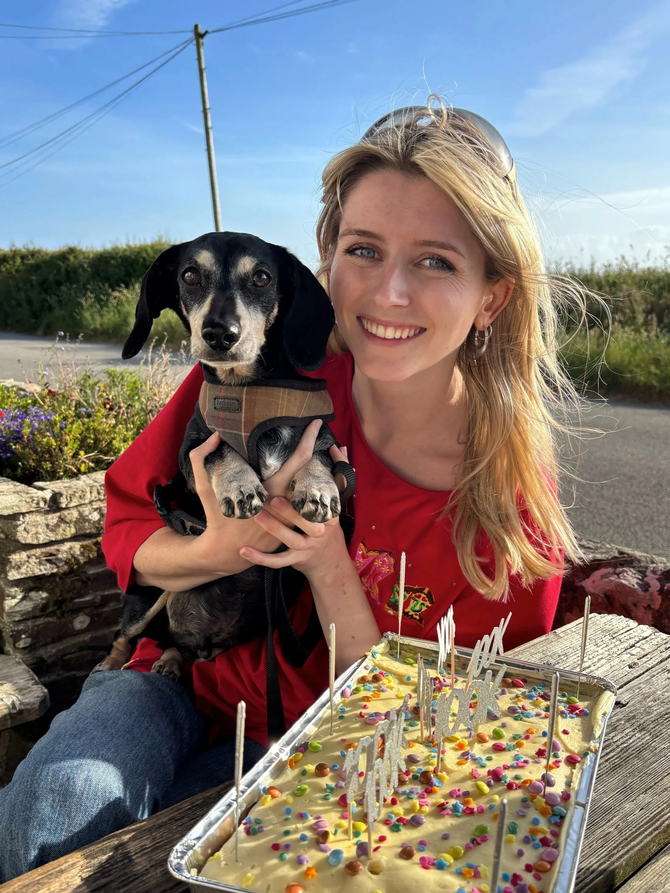 Evie, a young woman with blonde hair is holding Wilson, a black sausage dog, in her arms. She isitting outdoors near a wooden table with colorful cake with candles and sprinkles, on a sunny day with a blue sky and greenery in the background.