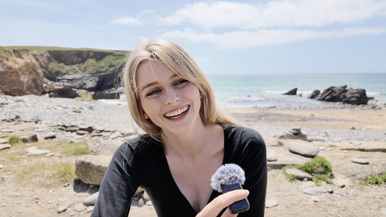 A headshot of Evie, a blonde woman and British English teacher, wears a black shirt and smiles while holding a microphone. She is on a sandy beach and the sea is blue.