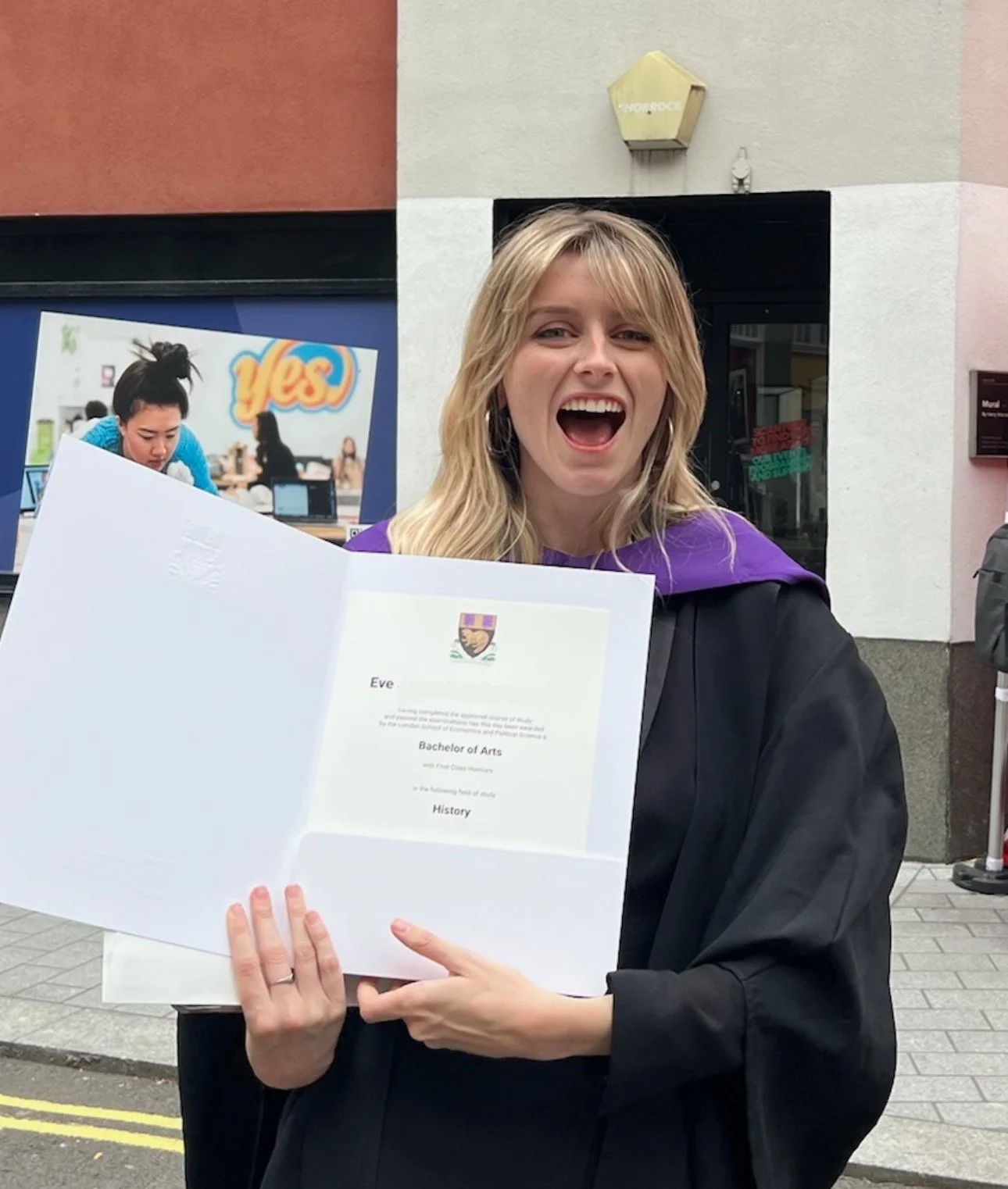 Evie stands on her graduation day in a black gown with a purple sash. She is holding her diploma from the London School of Economics and smiling widely.