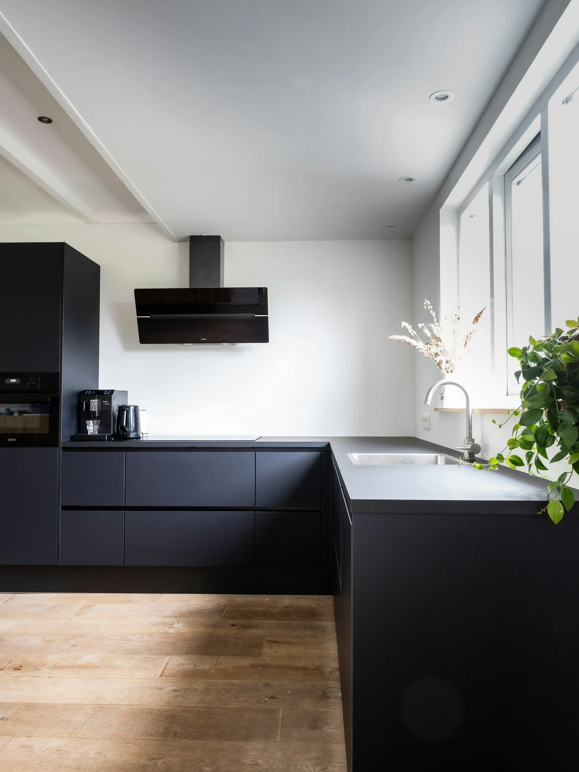 Modern kitchen with black cabinets, a black range hood, a sink near a window, and some green plants, with wooden floors.