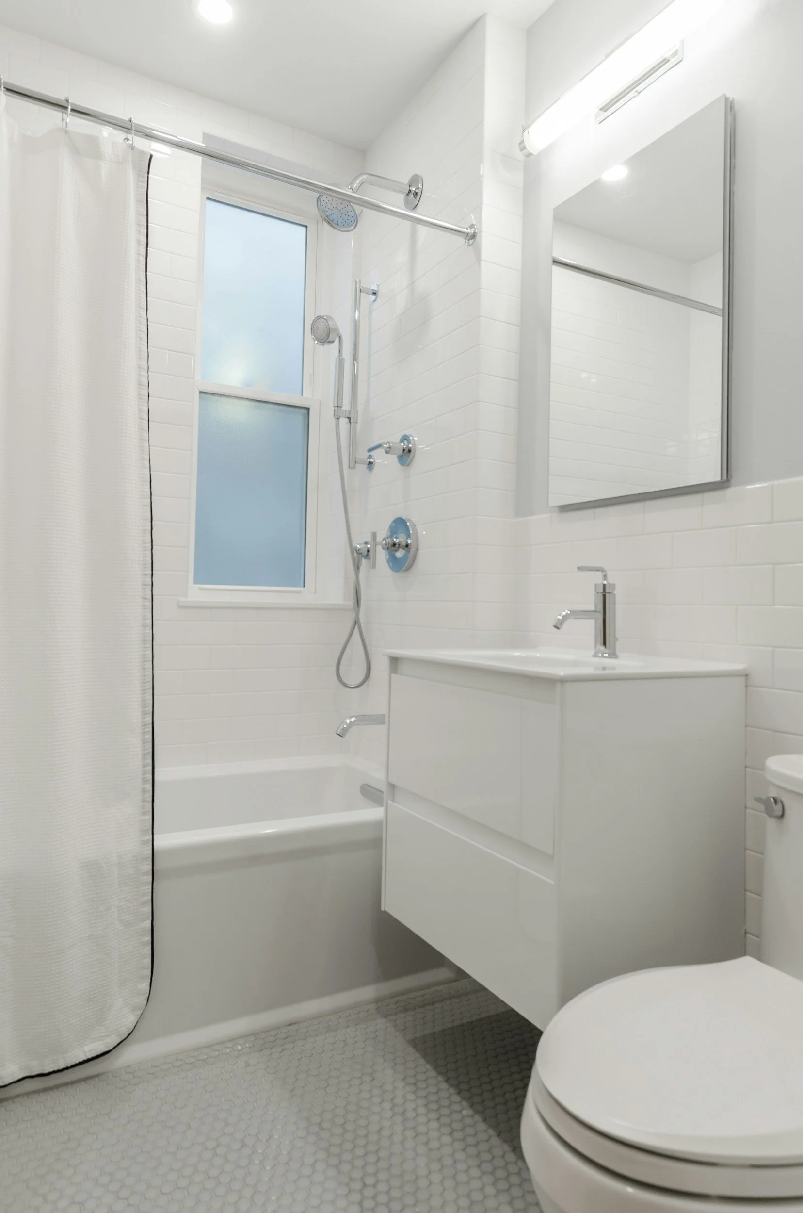 Modern small bathroom with a white tile shower, frosted window, white vanity with a sink, round toilet, and hexagon tile flooring.
