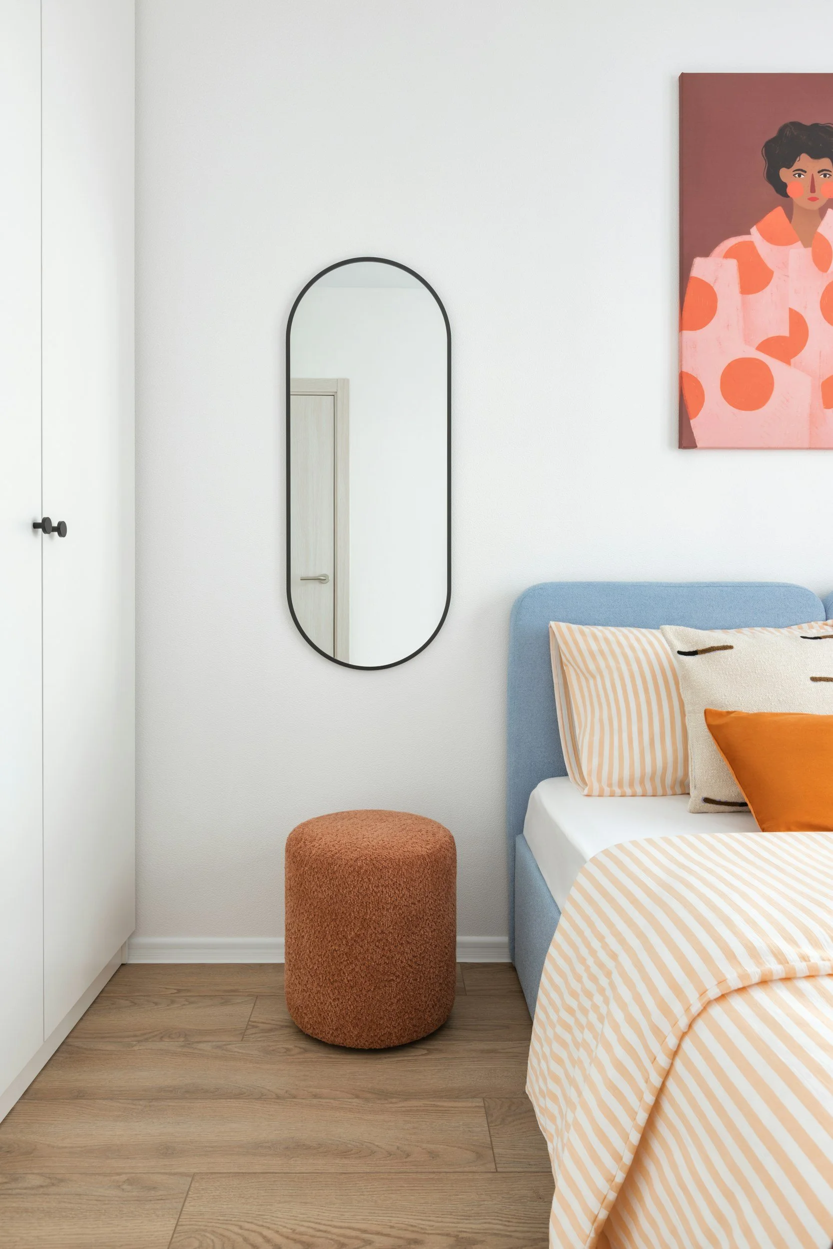 A bedroom corner with a bed featuring an orange and white striped duvet and pillows, a blue headboard, a small orange ottoman, a tall black-framed oval mirror on a white wall, and a colorful abstract portrait hanging above the bed.