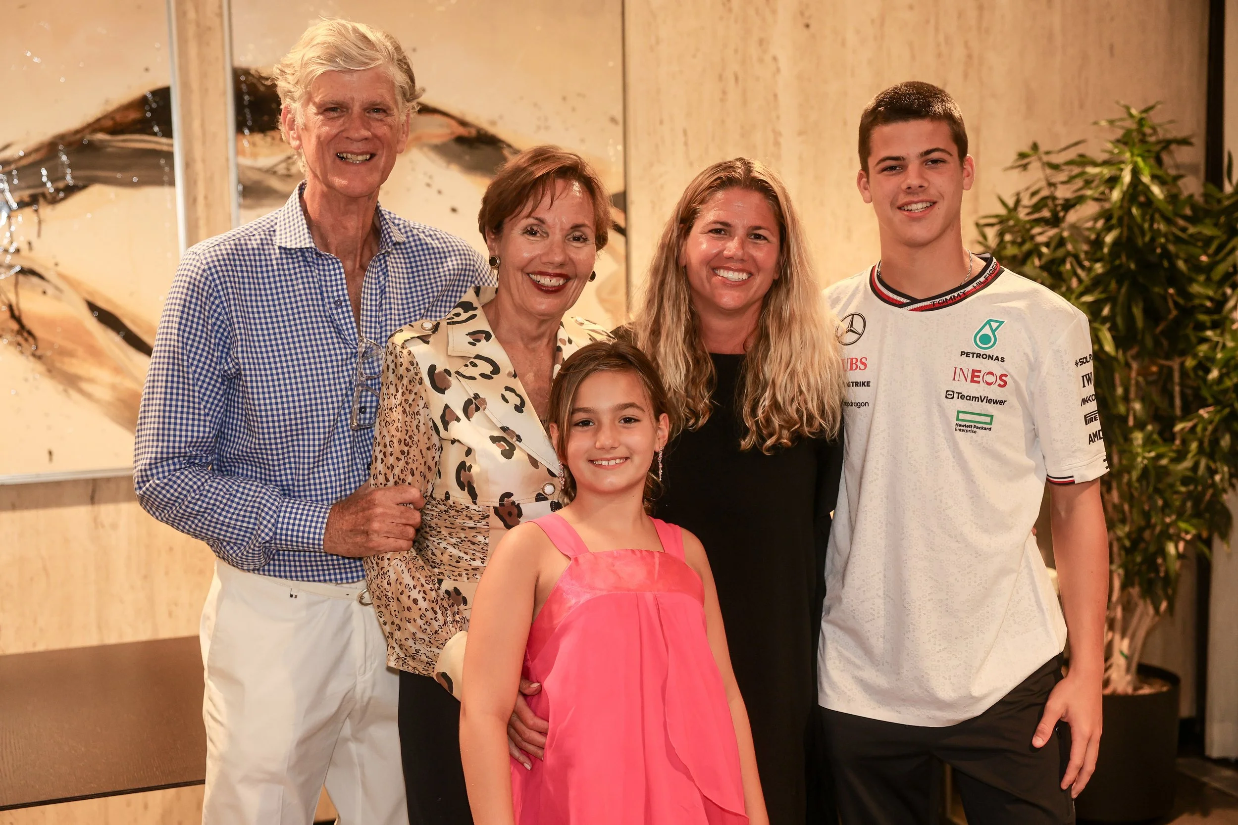 A group of five people, including an elderly man, an elderly woman, a young girl, a young woman, and a young man, smiling and standing close together indoors with a nature-themed abstract painting and greenery in the background.