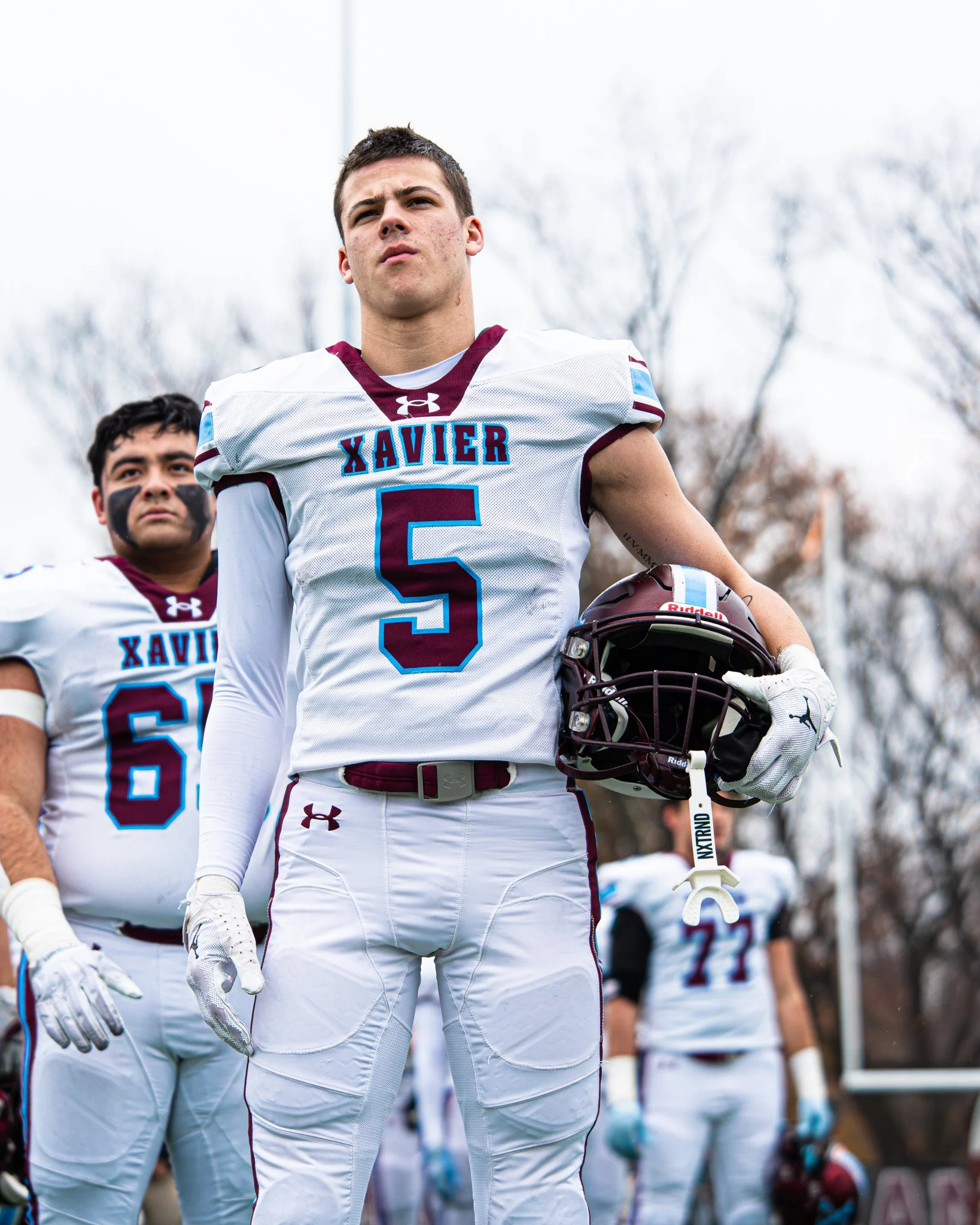 A young male football player in a white uniform with maroon and blue accents, holding a maroon helmet and standing with other players on a football field, looking determined amidst an overcast sky and barren trees.