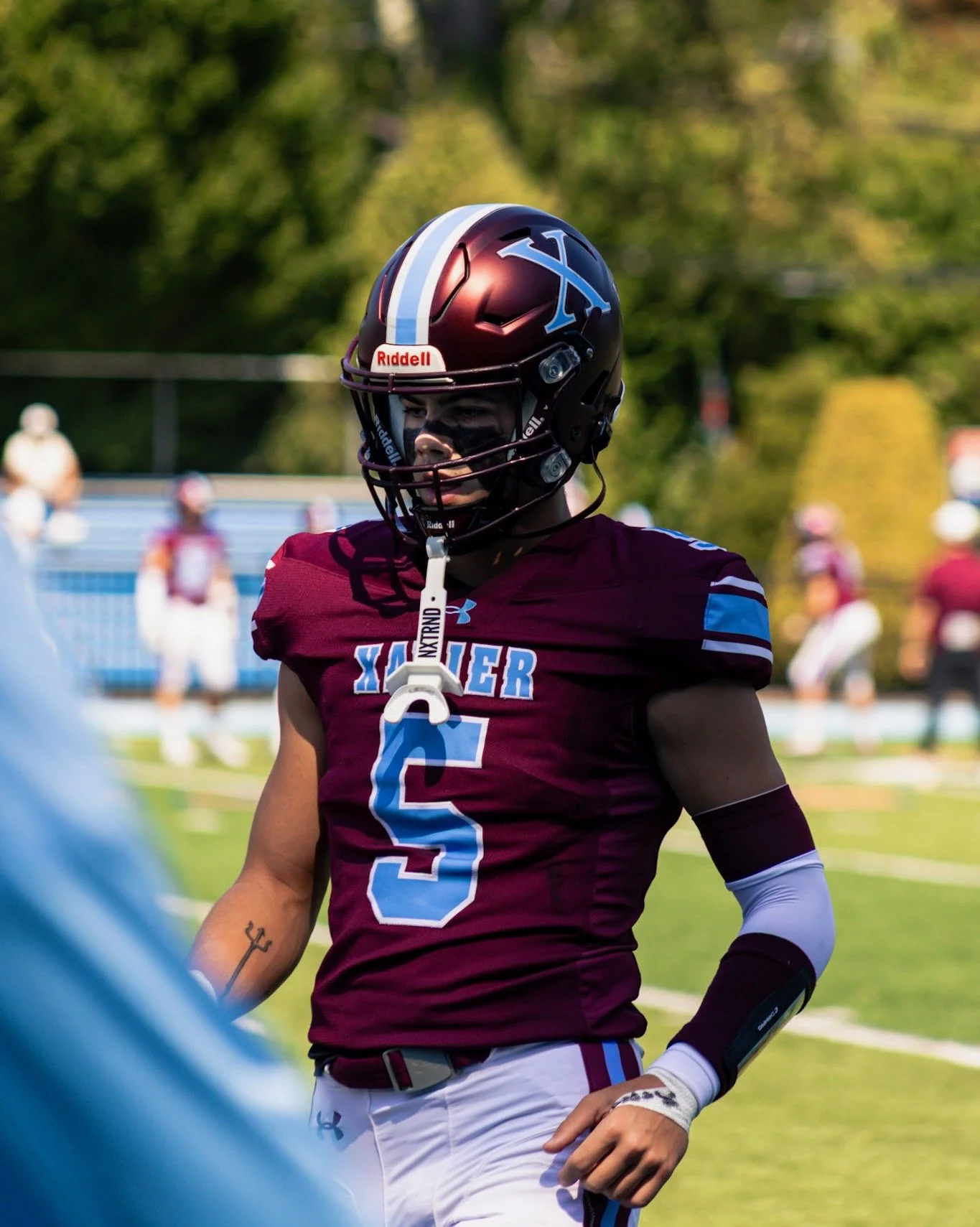 An American football player wearing a maroon and blue uniform with the number 5 and a helmet with a large X on it, standing on a football field during a game.