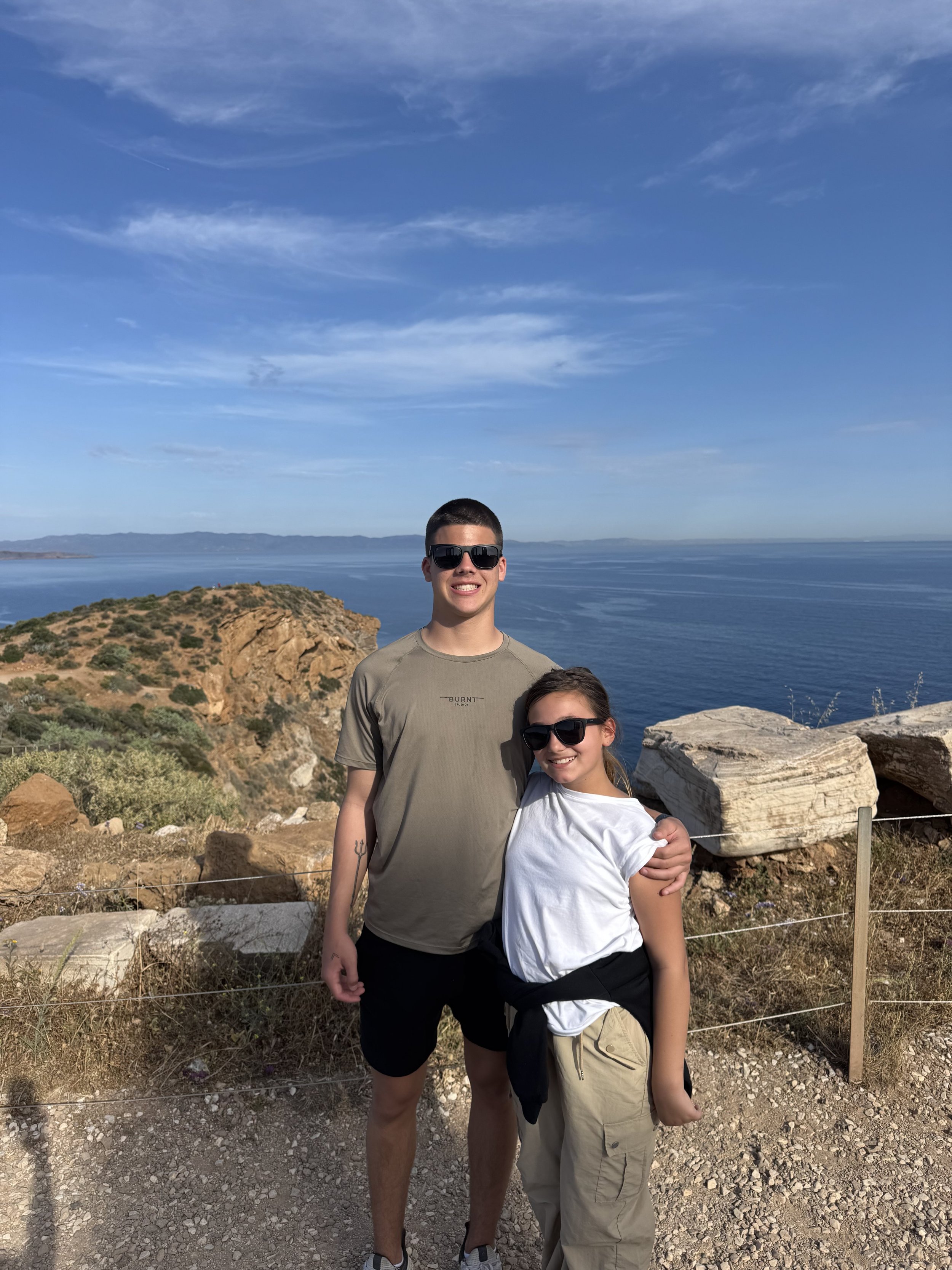 Two young people, a man and a girl, standing side by side outdoors near the coast with hills, rocks, and water in the background, both wearing sunglasses and smiling.
