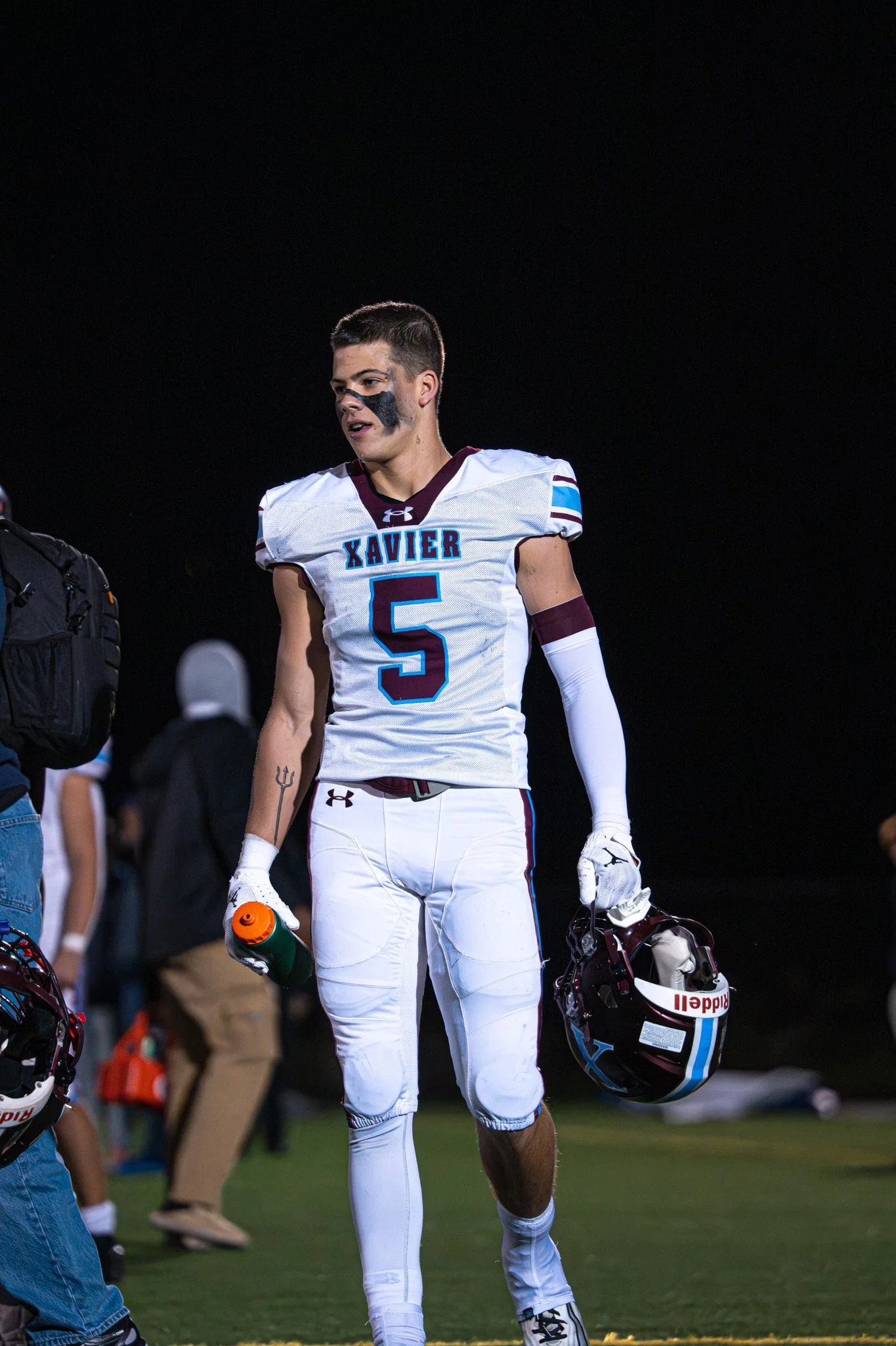 A high school football player in a white uniform with blue and maroon accents, holding a helmet in one hand and a water bottle in the other, walking on the field at night.