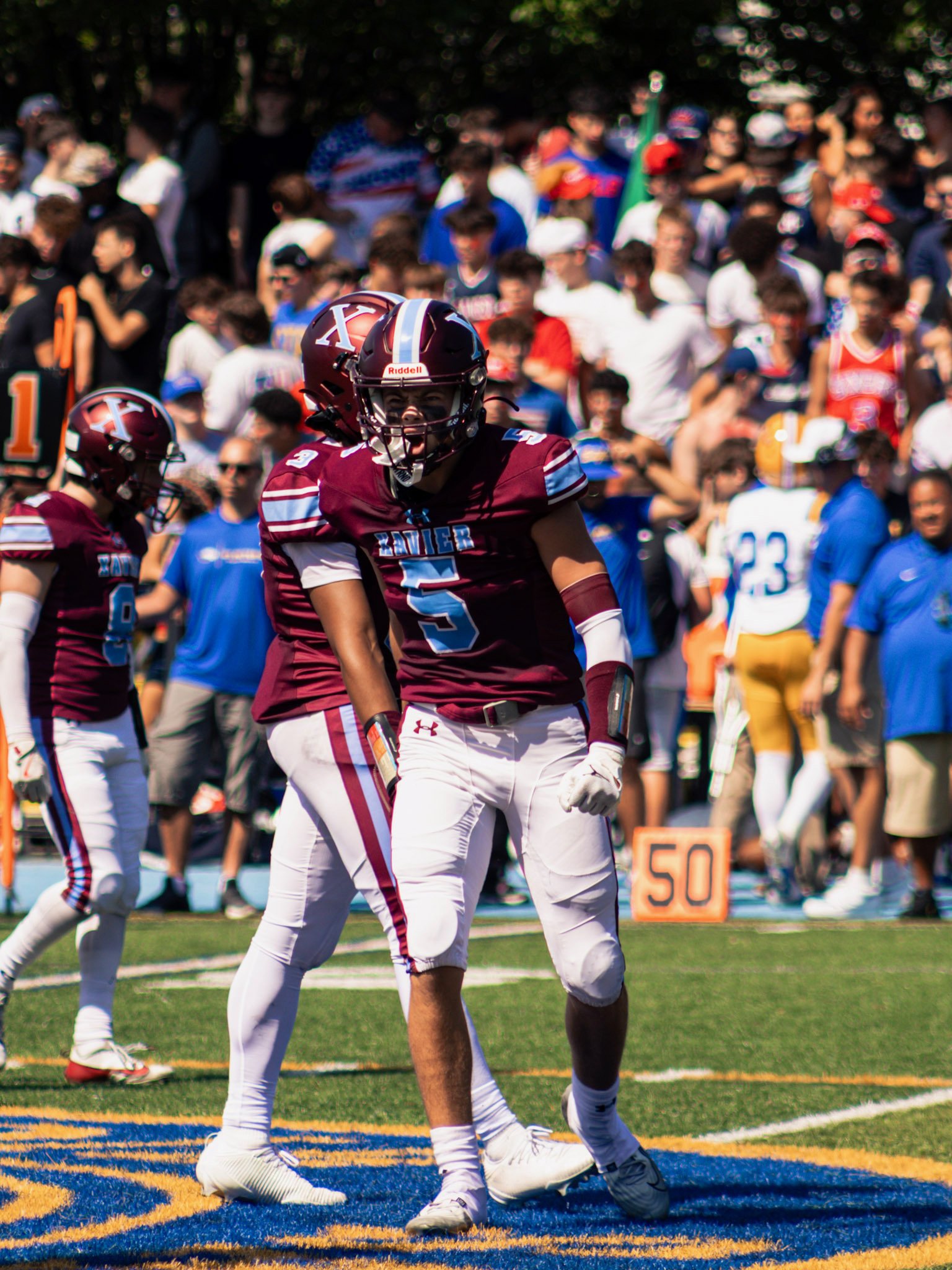 American football game with players in maroon and white uniforms on a field with orange and blue end zone. One player in maroon jersey number 5 appears to be celebrating or celebrating a play. Spectators in the background watch the game.