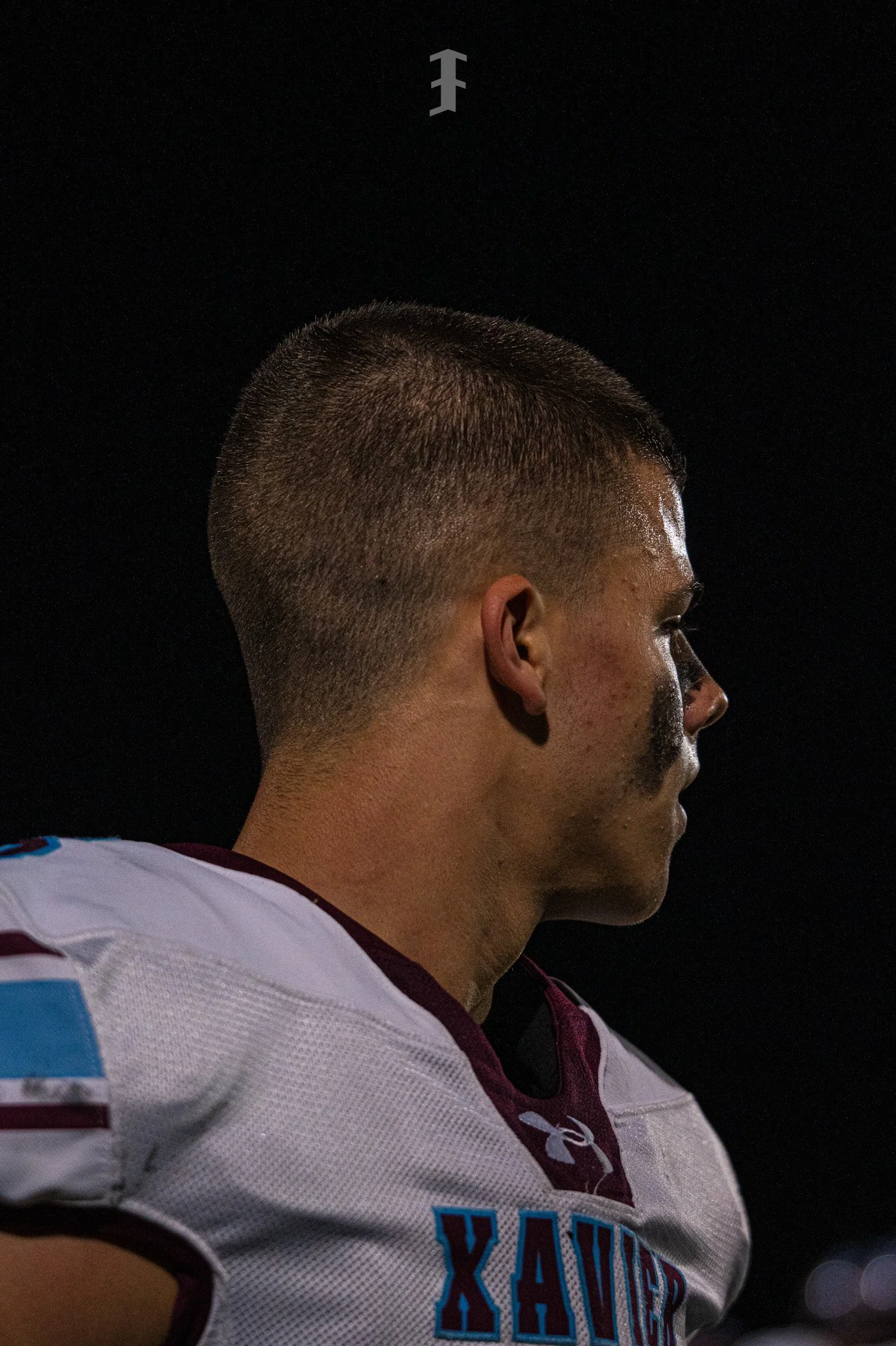 A close-up side profile of a male athlete with a short haircut, wearing a white sports jersey with maroon and blue accents, and a black background.