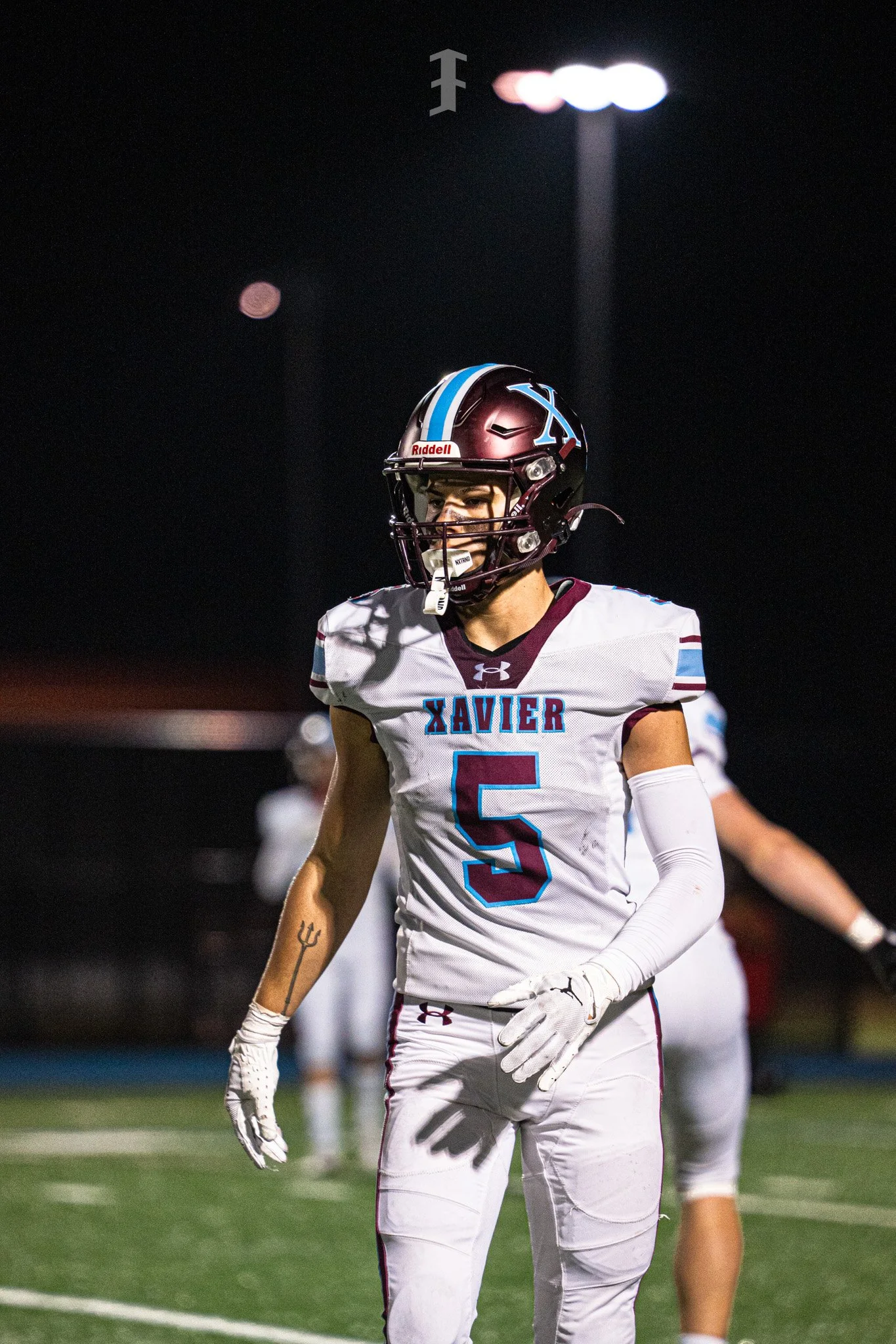 A football player from Xavier wearing a white jersey with the number 5, a maroon helmet with a blue stripe, and white gloves stands on the field at night under stadium lights.