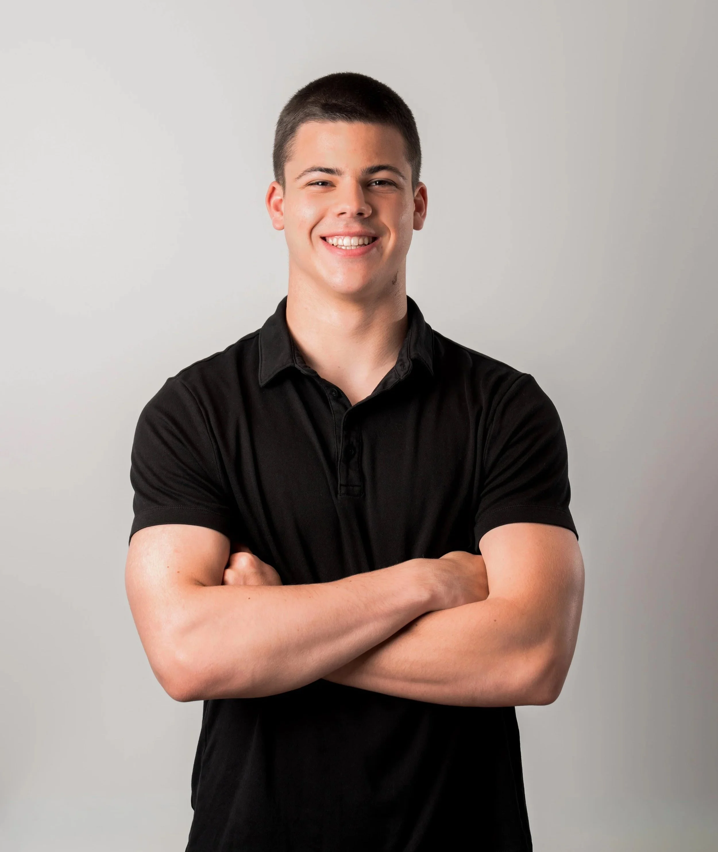 A young man with short dark hair, wearing a black short-sleeve shirt, smiling with arms crossed against a plain light gray background.