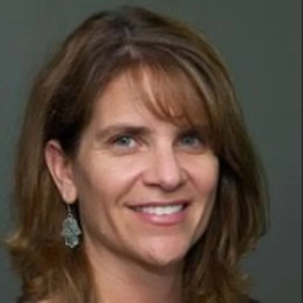 A smiling woman with shoulder-length brown hair and blue eyes, wearing earrings against a gray background.